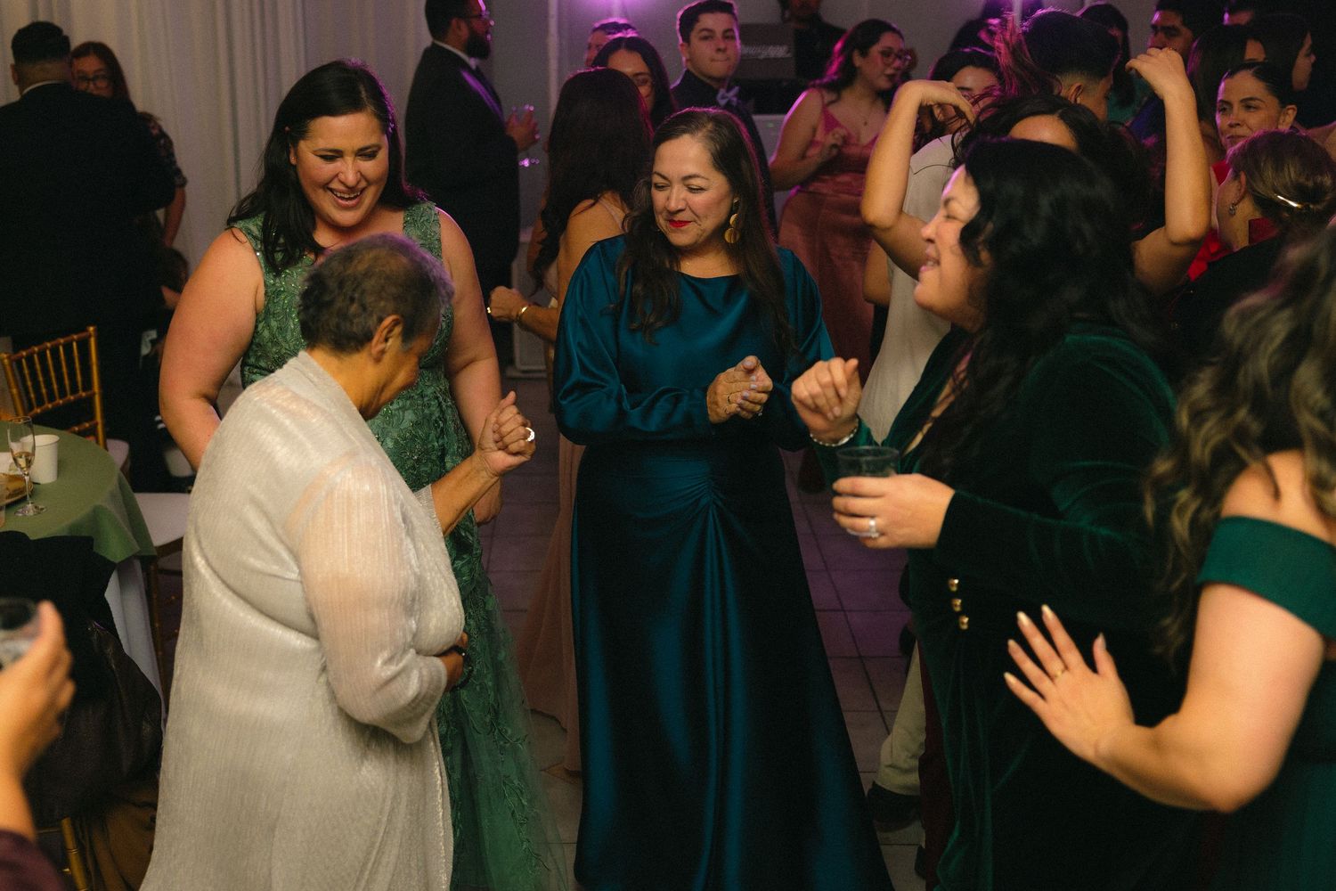 Wedding guests in dark blue dresses dance and celebrate at an indoor reception with purple lighting.
