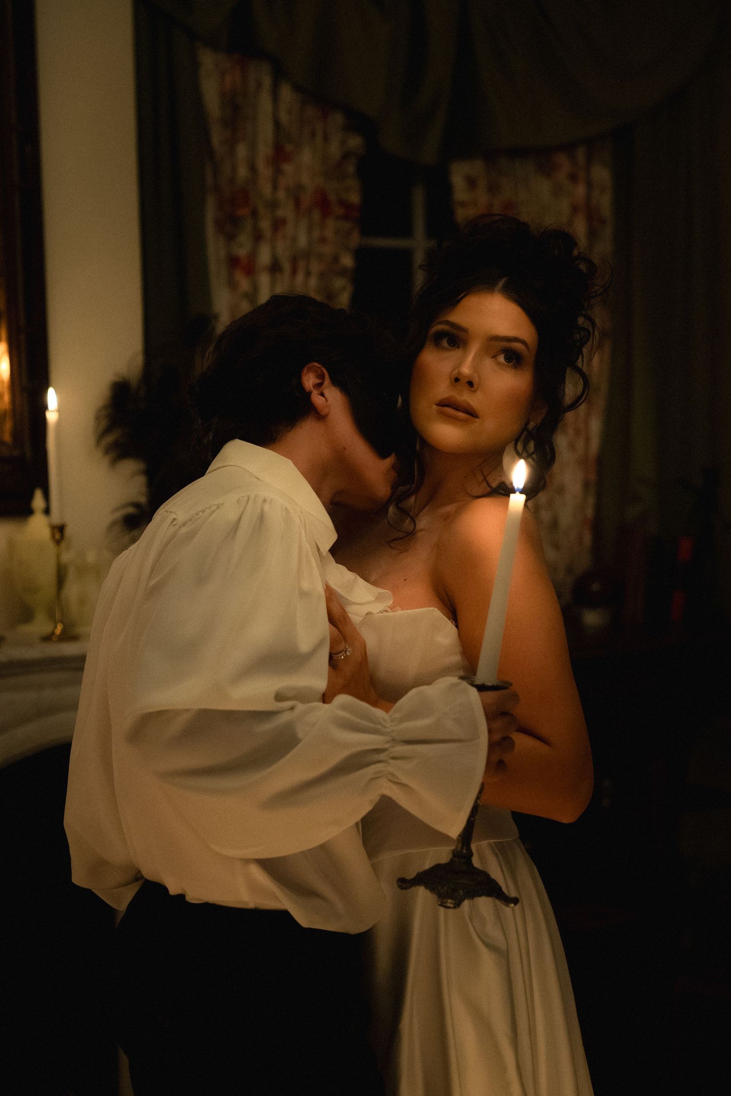 A couple in white attire dancing intimately in a dimly lit room with curtains visible in the background.