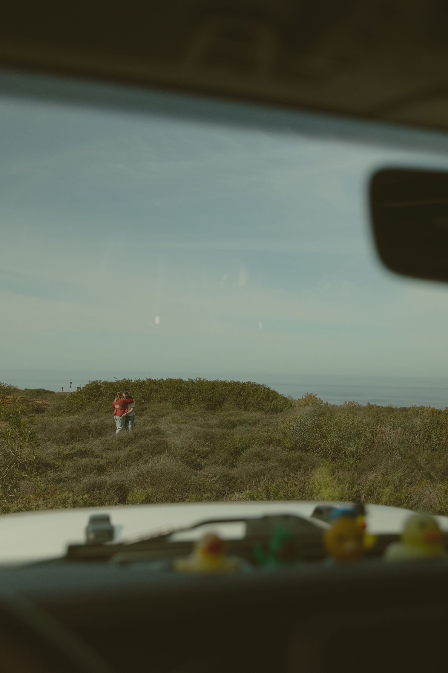 A distant figure stands on a hilltop viewed through a car windshield with dashboard decorations.
