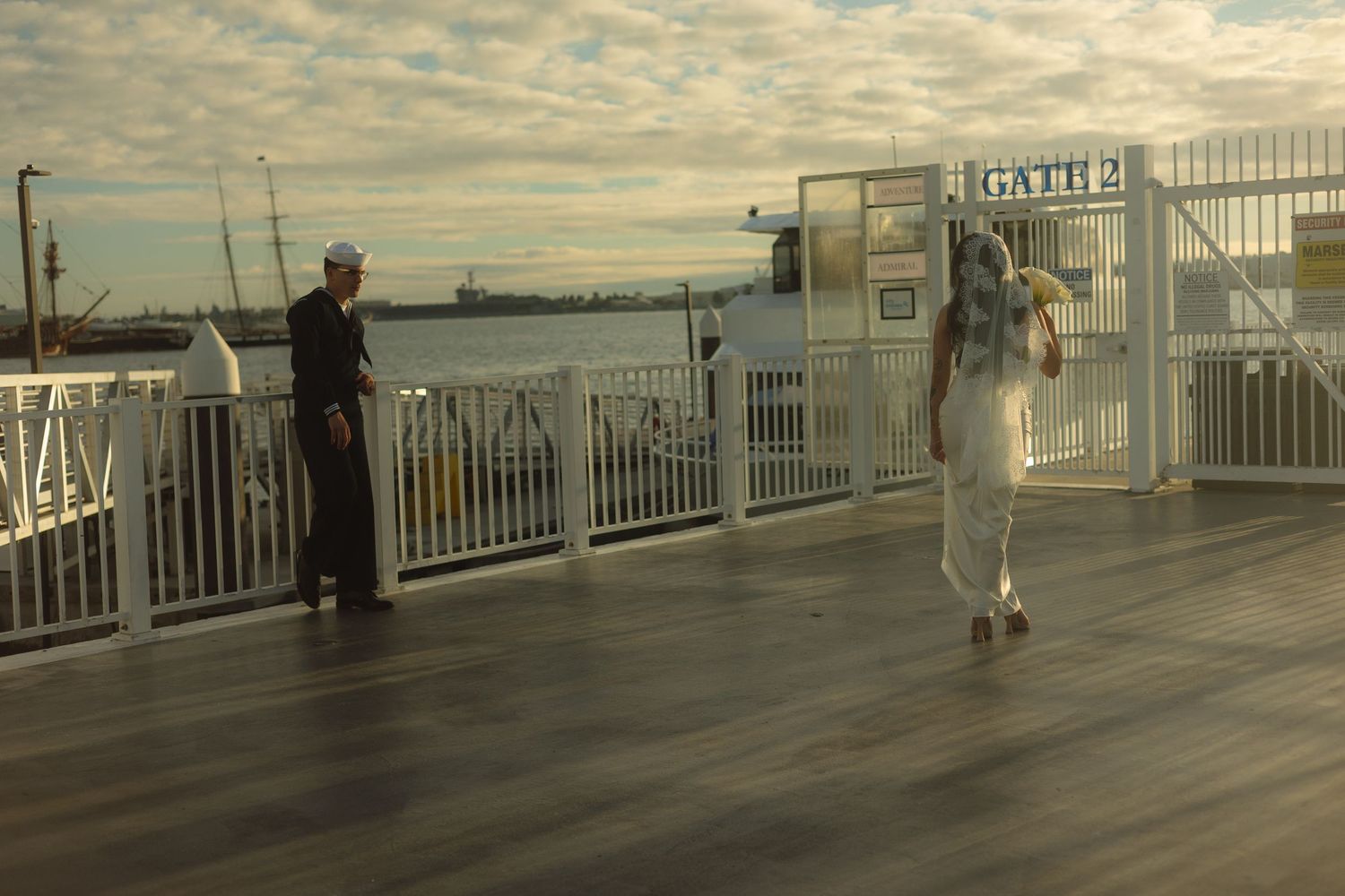 People stand on a wooden deck by railing overlooking water at sunset with atmospheric lighting.