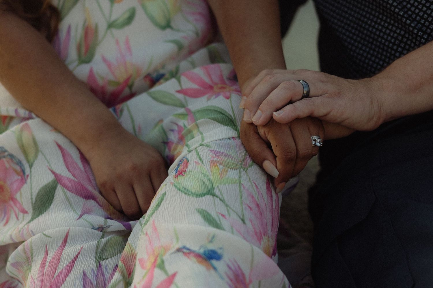 Two hands rest together on floral patterned fabric, with one wearing a sparkly engagement ring.