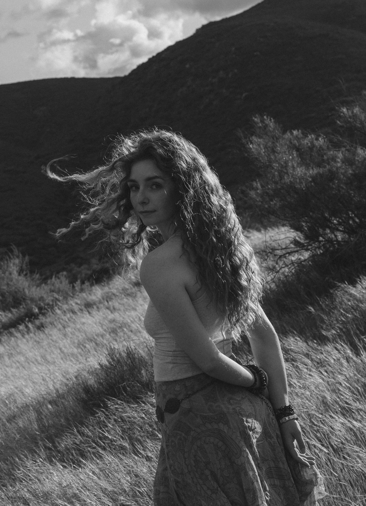 A black and white artistic portrait of a figure with flowing curly hair standing on a hillside against a dramatic sky.