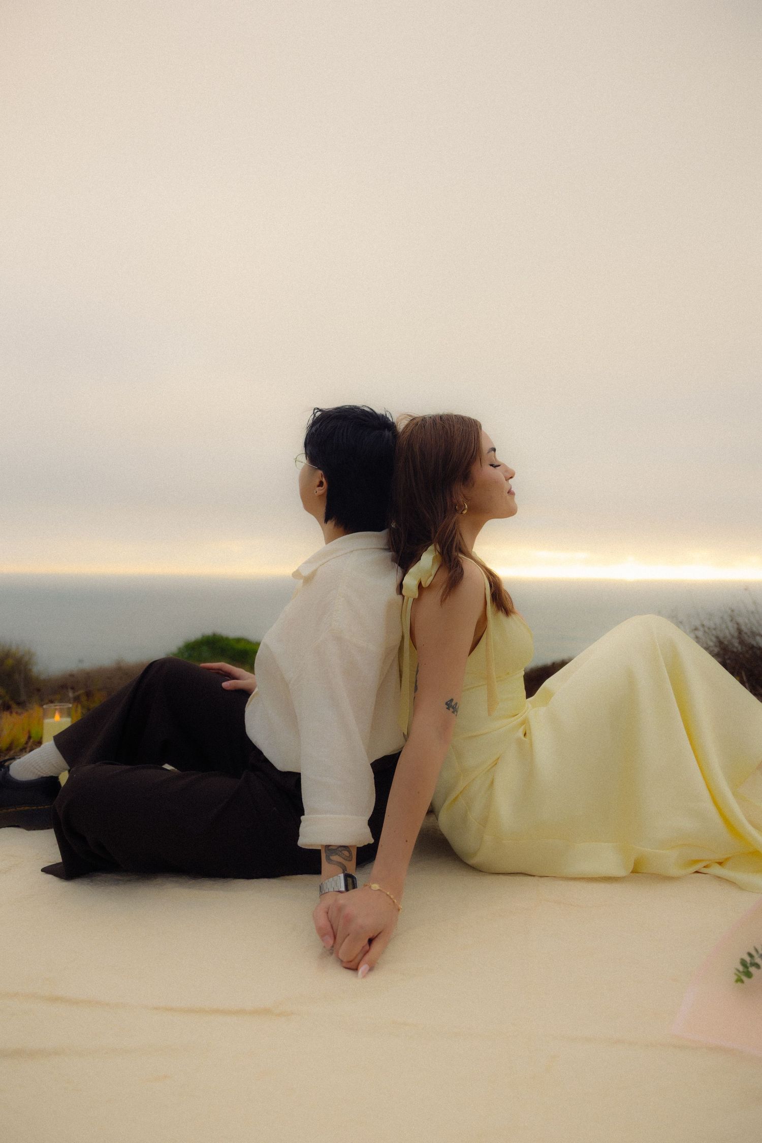 Two people in formal attire sit back-to-back on a light surface at sunset overlooking an ocean view.