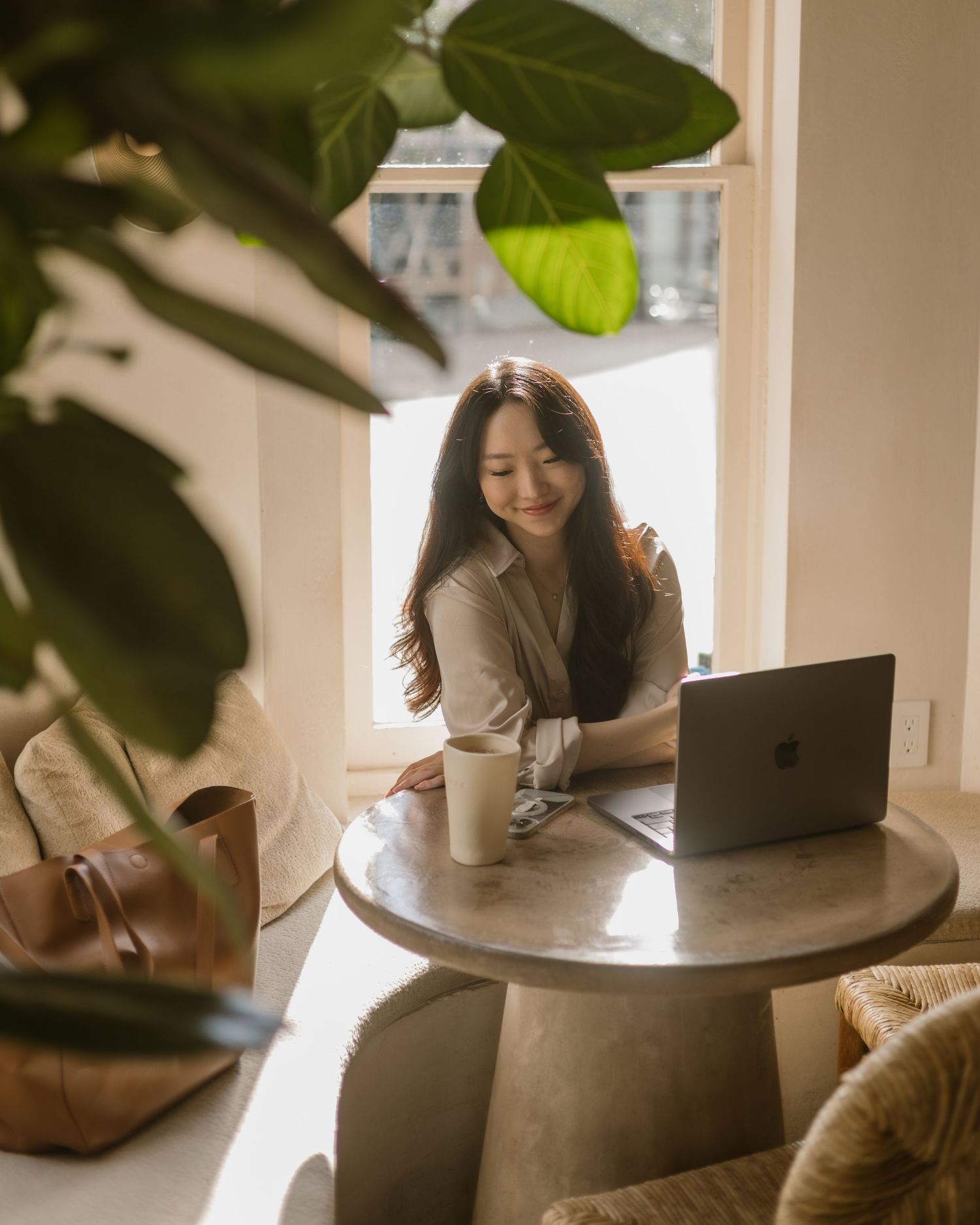 Someone works on a laptop at a round table with coffee in a bright room with houseplants and natural lighting.