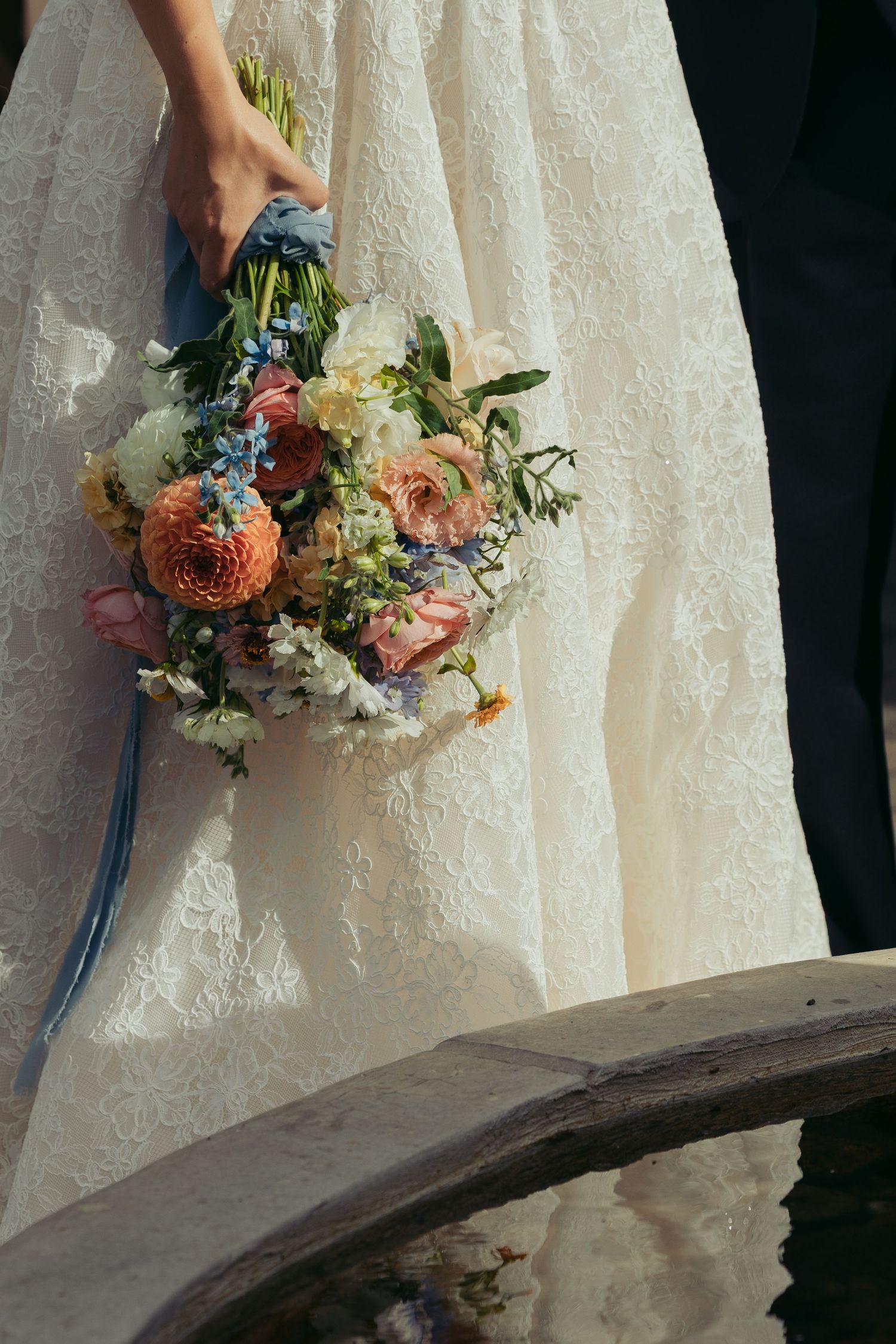 Bride in lace wedding gown holding a colorful bouquet of pink, blue, and white flowers near a wooden well.