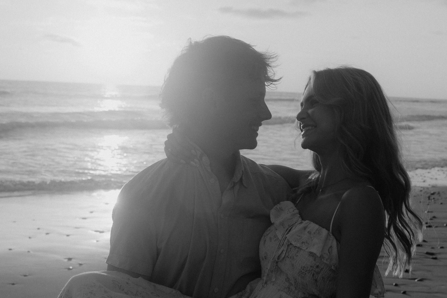 Silhouettes of couple sitting on beach at sunset in black and white.
