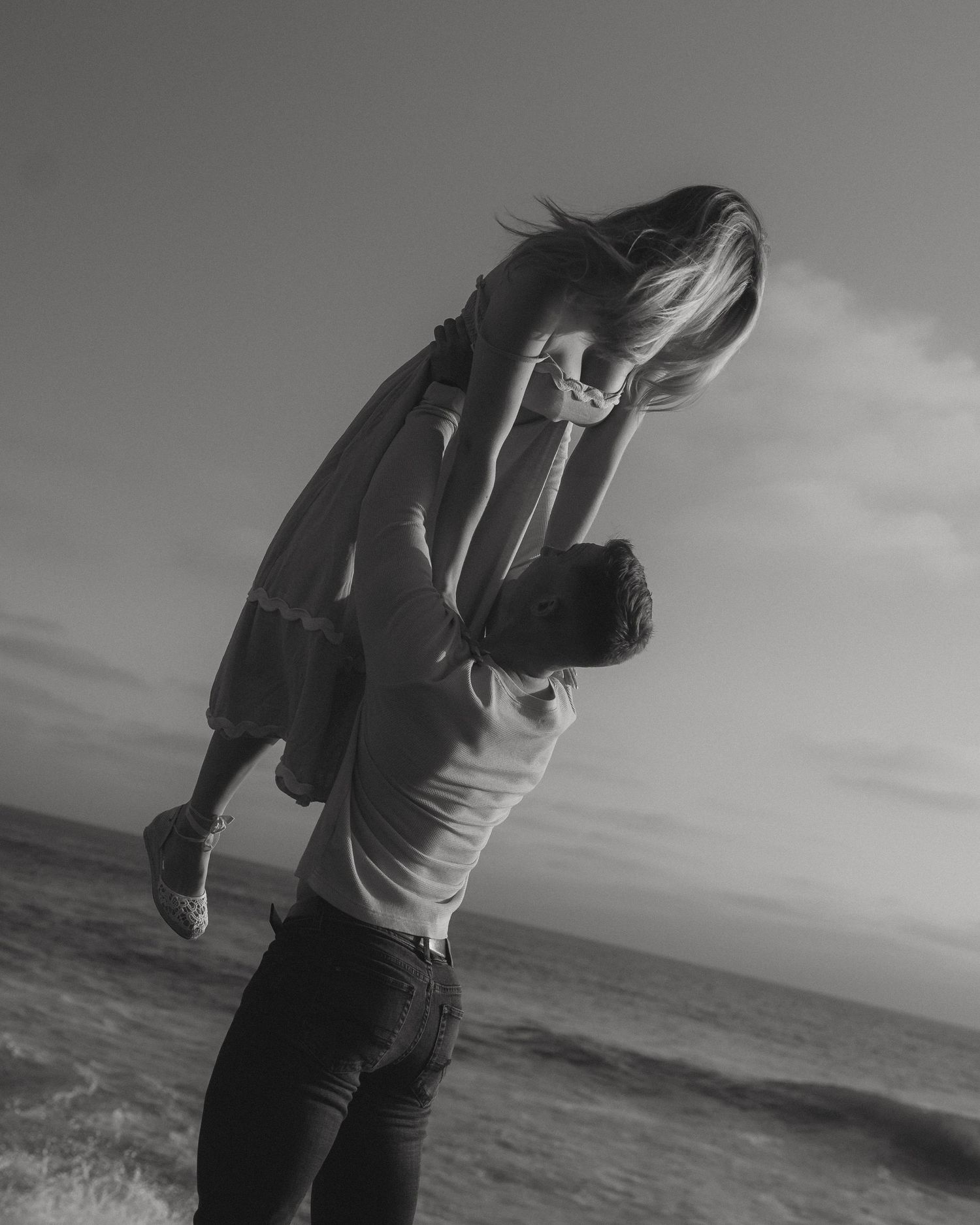 Black and white photo series of a couple embracing on a beach with ocean waves and cloudy sky in the background.