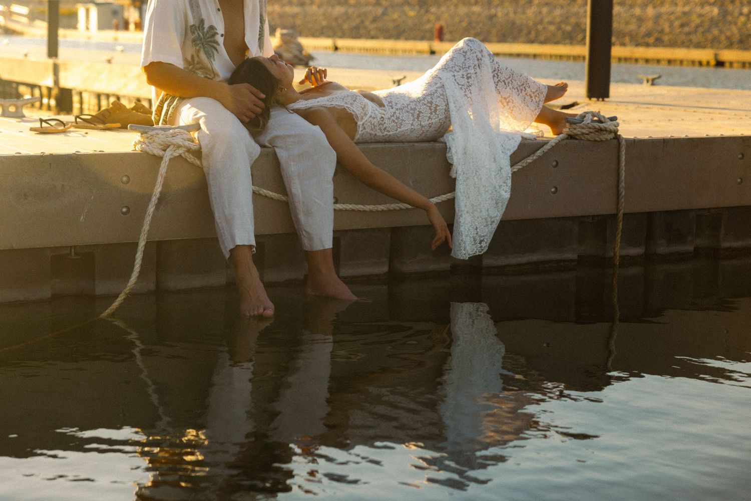 People's legs dangle over a dock at sunset as they sit together enjoying the peaceful waterfront view.