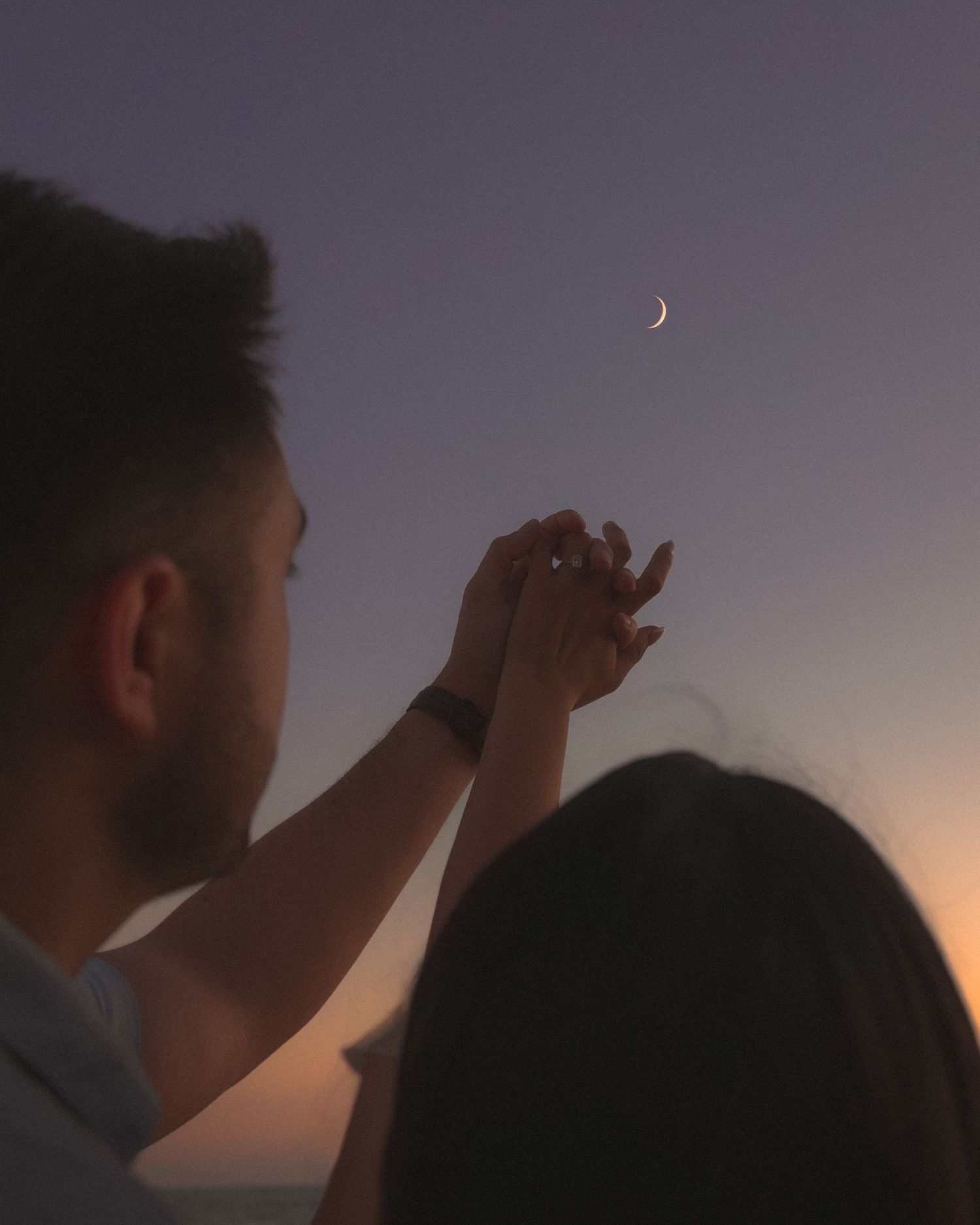 A silhouetted figure holds their hand up to the crescent moon in the dusky purple sky.