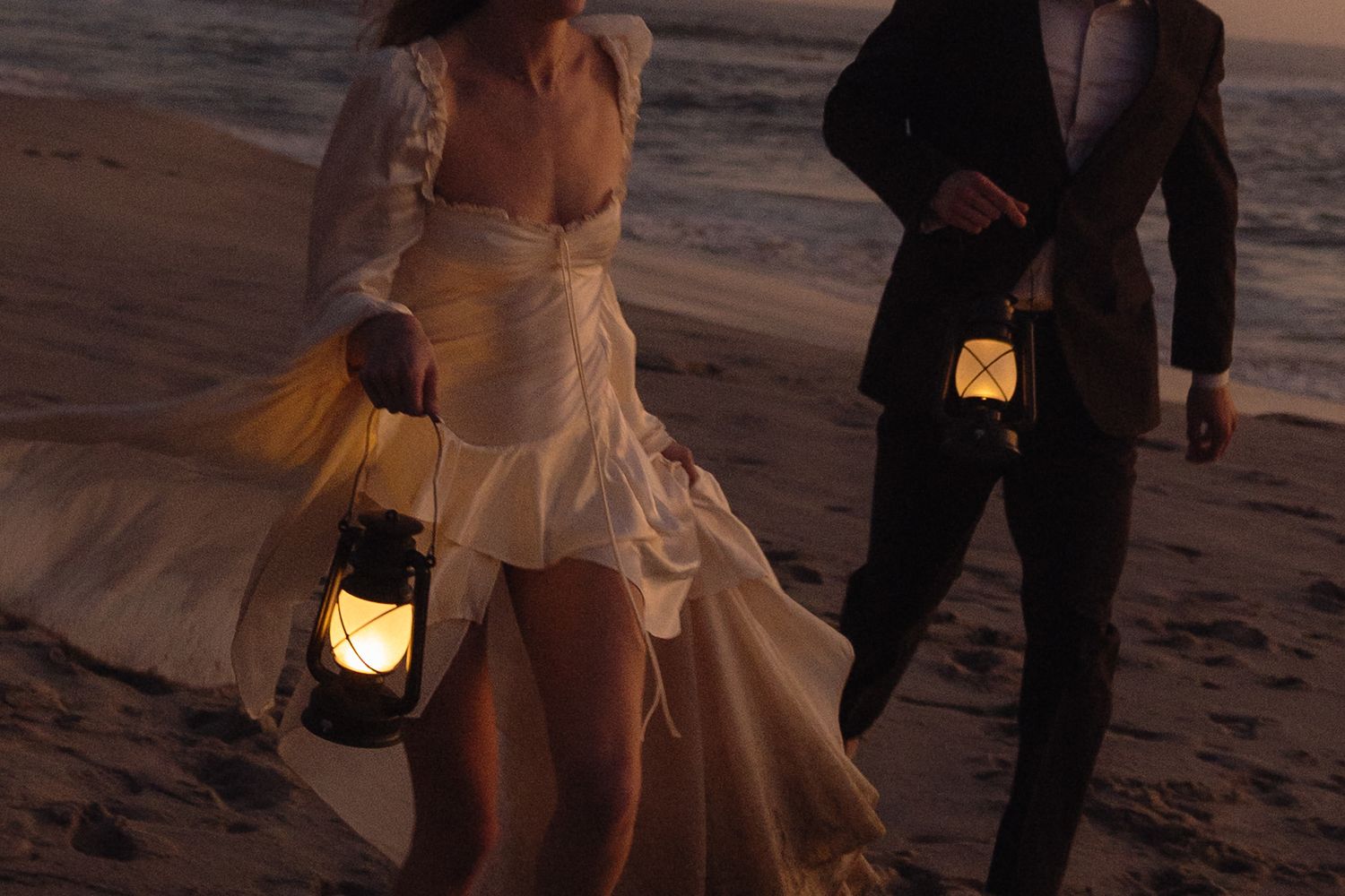 Bride and groom walking on beach at sunset holding glowing lanterns, romantic wedding photography.