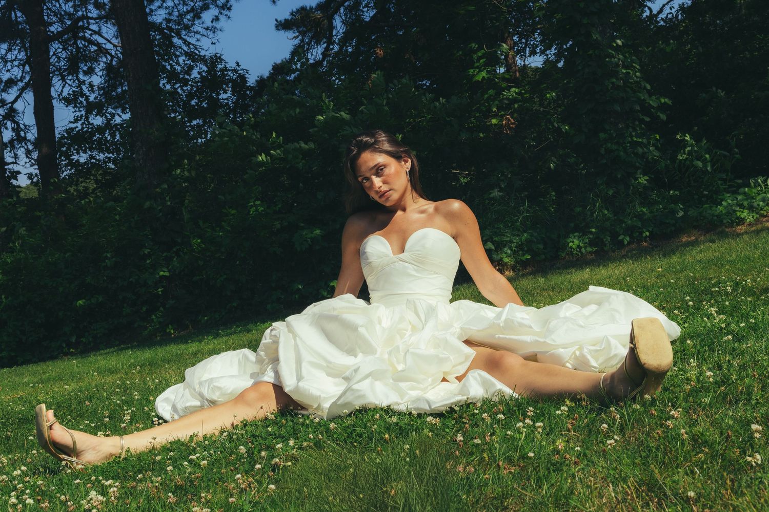 Barefoot bride in white strapless gown sitting on green grass in sunny park.