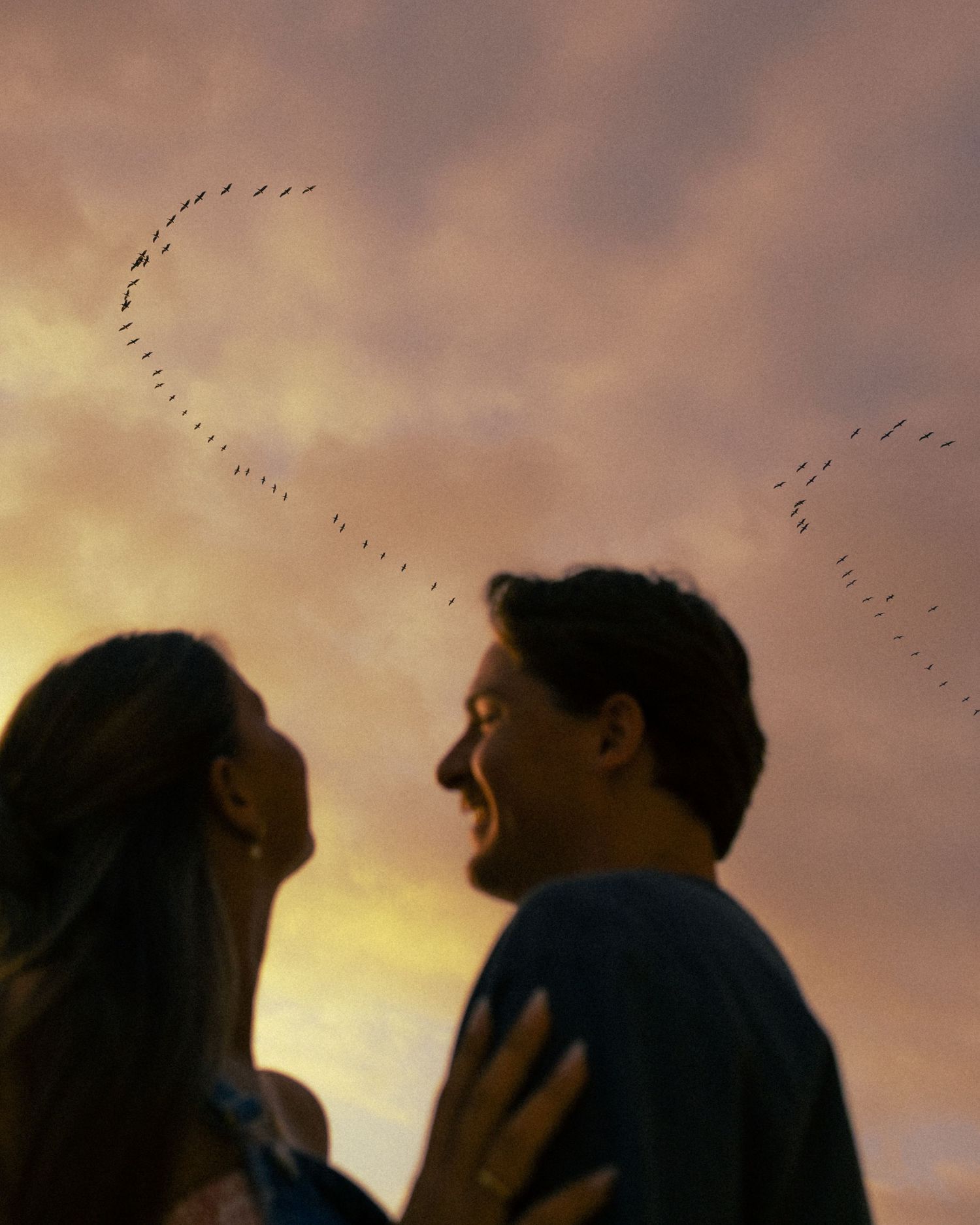 Couple sharing a romantic moment at sunset with birds forming a heart shape in the colorful sky above them.