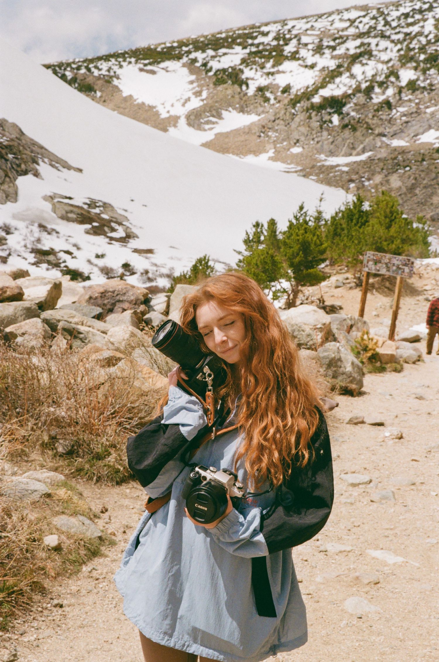 A redhead woman holding two cameras on a snowy mountain trail, wearing a light blue dress and black jacket.