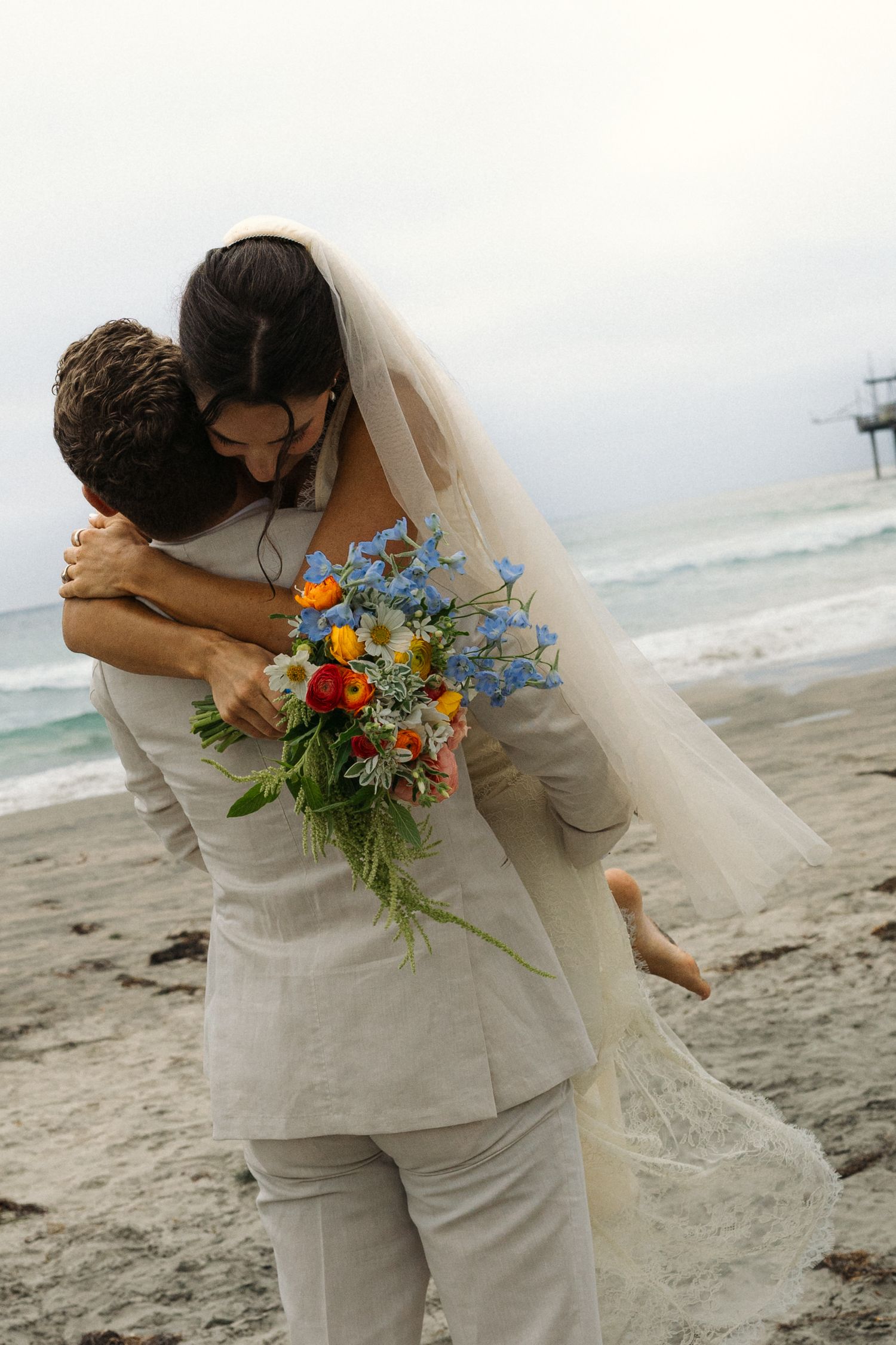 Groom lifting bride in wedding dress on beach, sharing romantic embrace with colorful bouquet, ocean in background.