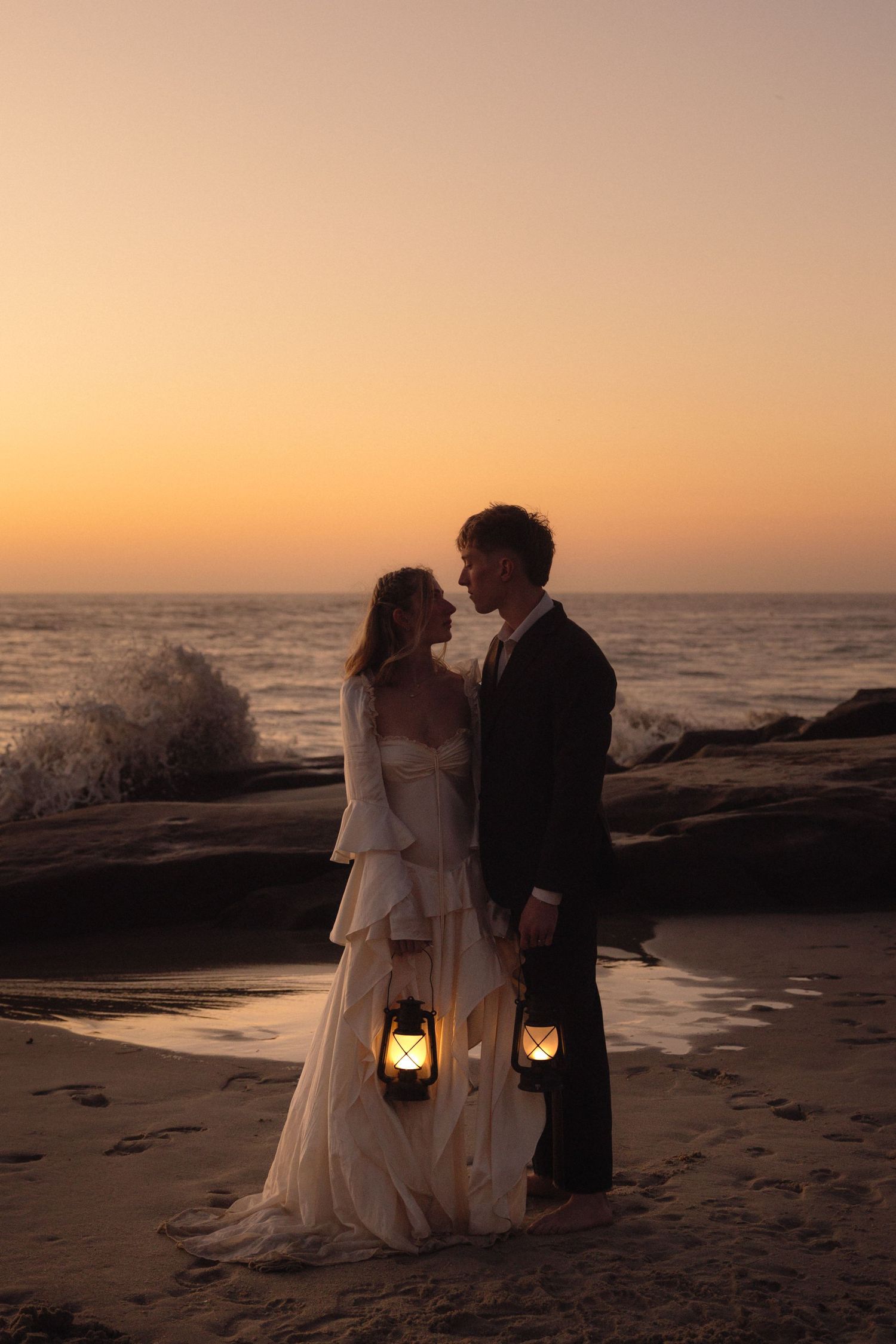 A couple in wedding attire stands on a beach at sunset holding lanterns against an orange sky and crashing waves.