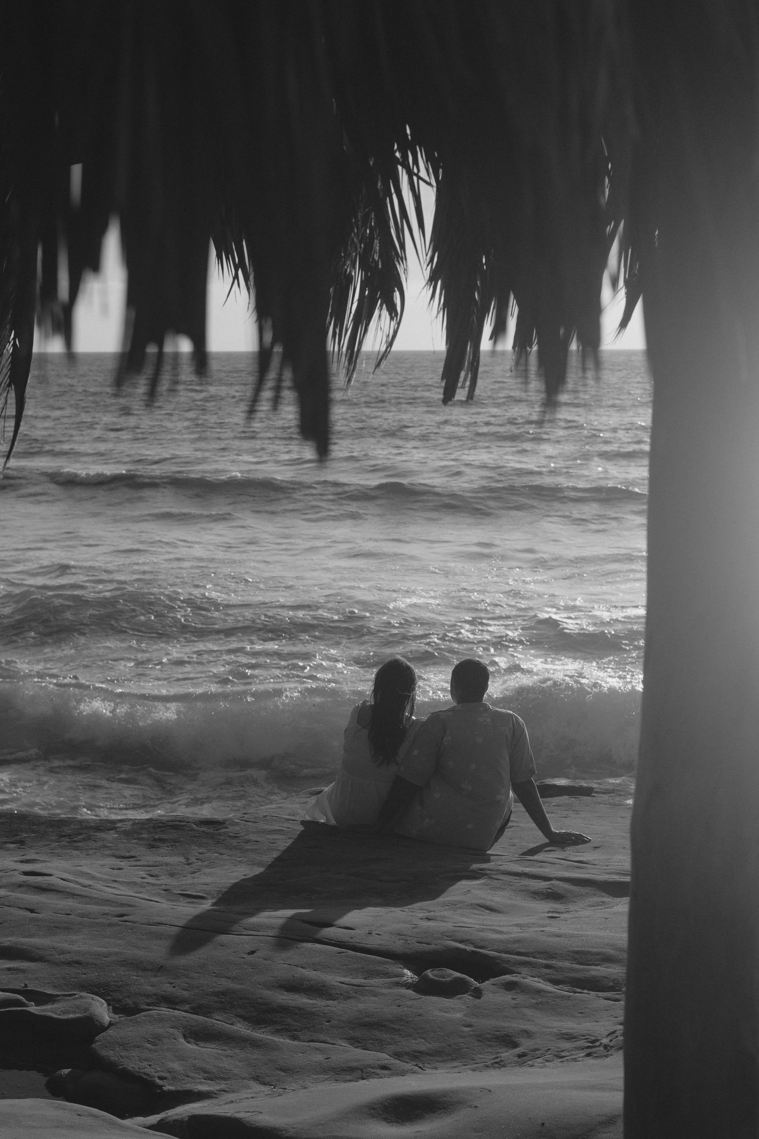 A couple sits on the beach watching the ocean waves beneath palm fronds in a romantic black and white scene.