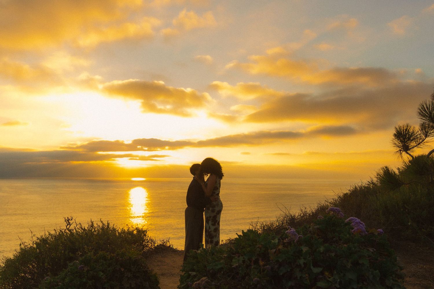 A scenic oceanfront wedding ceremony silhouette against a dramatic orange and gold sunset sky.