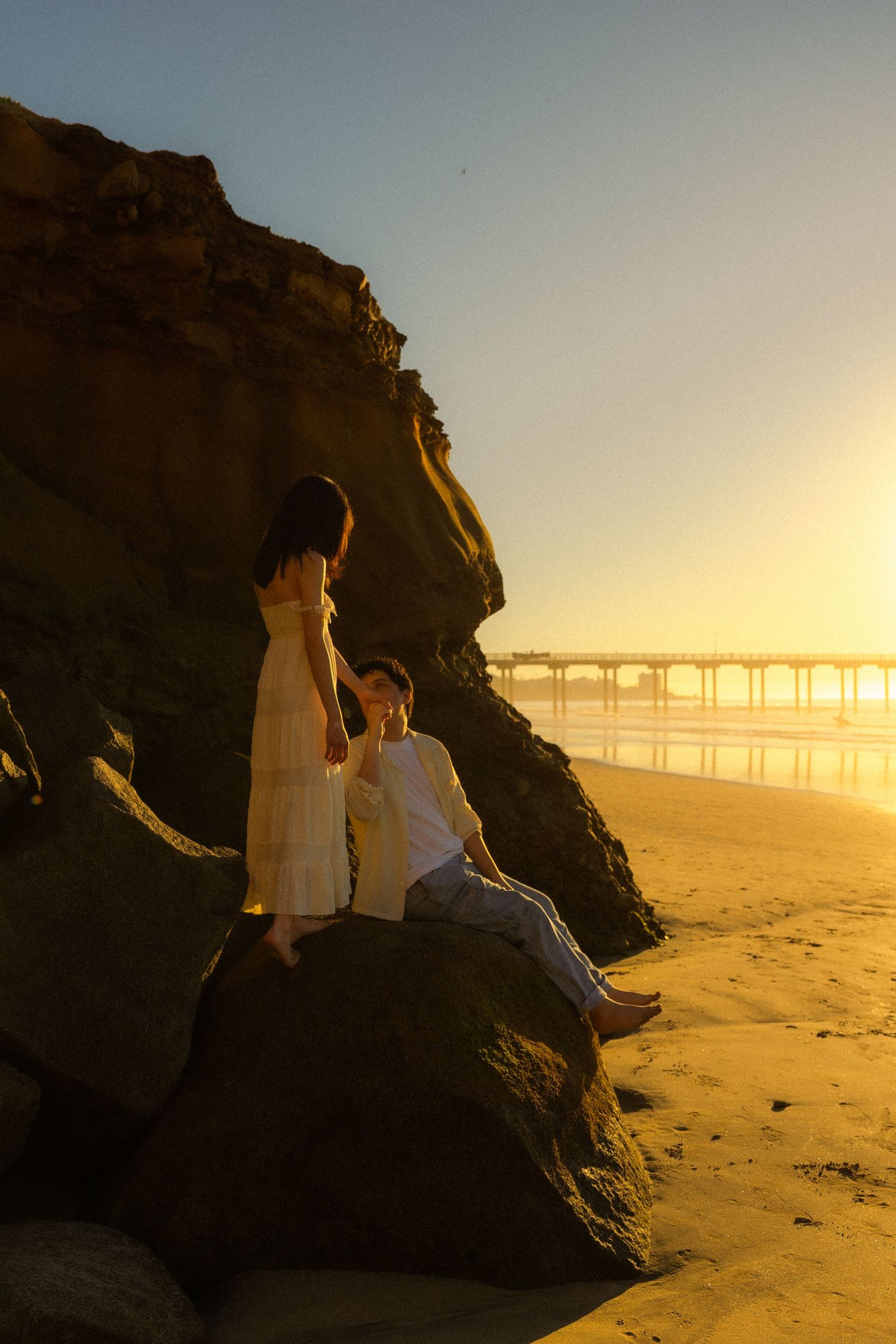 A couple sits together on coastal rocks at sunset with wind turbines visible in the golden distance.
