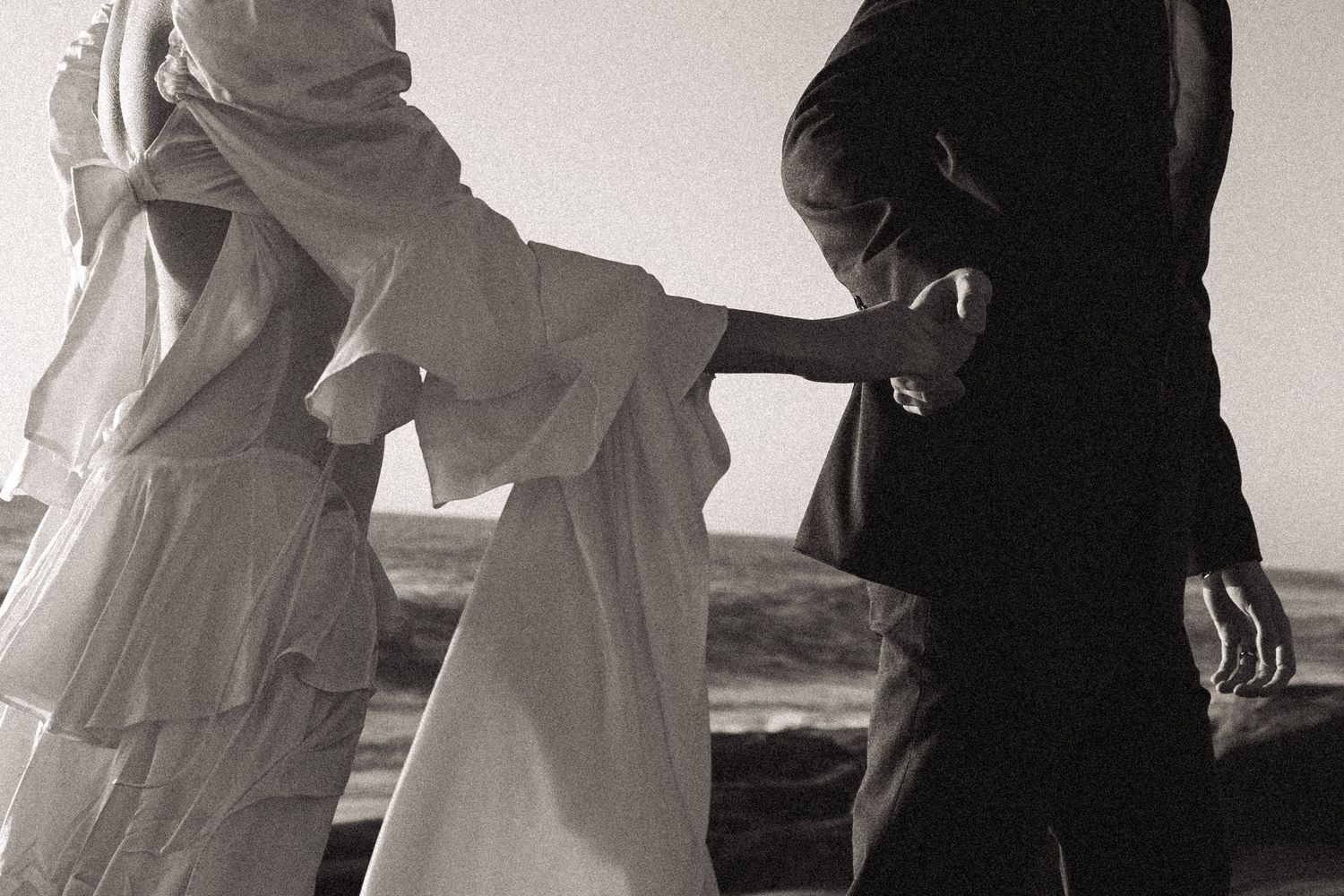 A couple holds hands on a beach with the bride wearing a flowing white tiered dress in black and white photography.