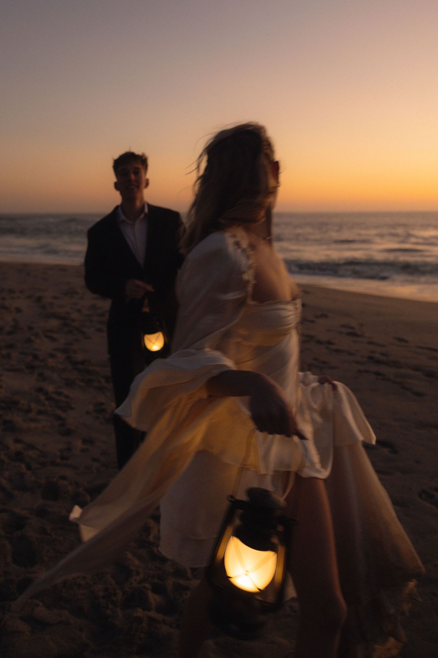 A romantic beach wedding photoshoot at sunset with glowing lanterns casting warm light on the sand.