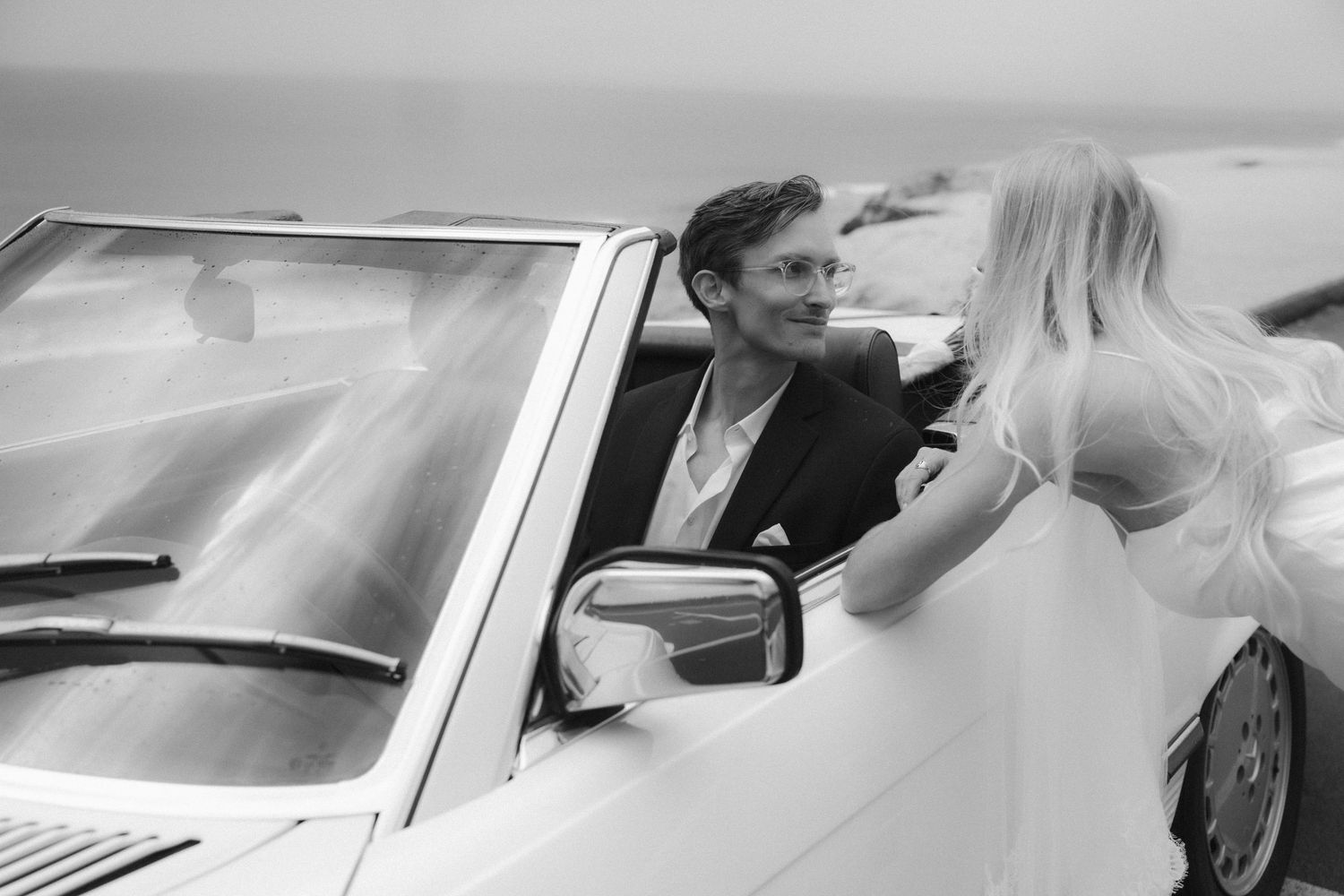 Black and white photo of a groom in a suit sitting in a white convertible car, smiling at a blonde bride.