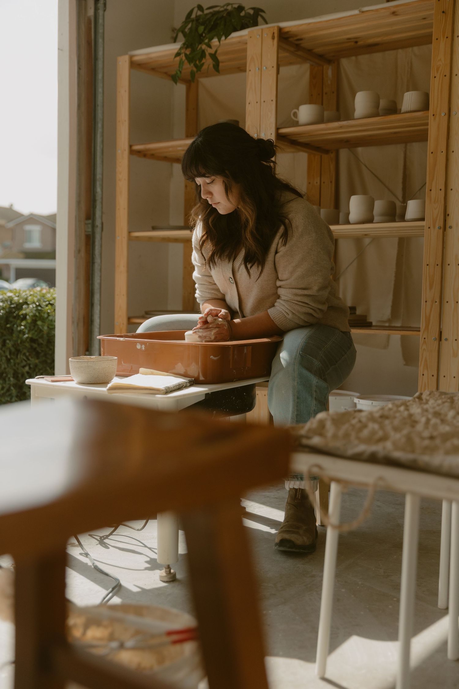 Person in beige sweater working at pottery studio with wooden shelving.