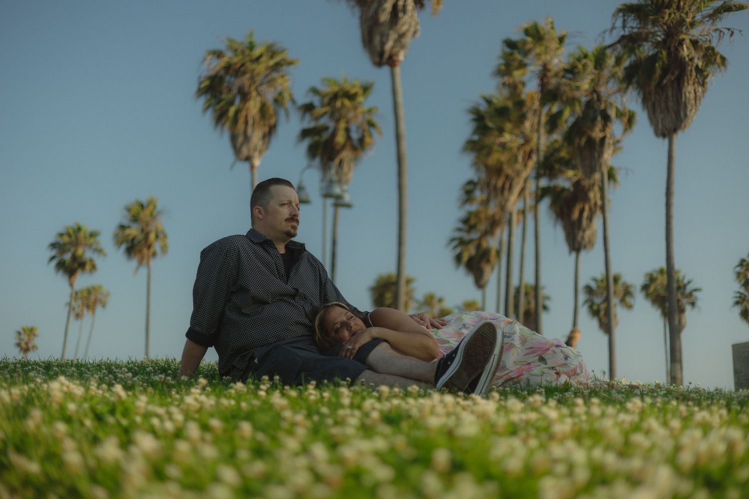Person relaxing on grass with tall palm trees towering against a bright blue sky in California.