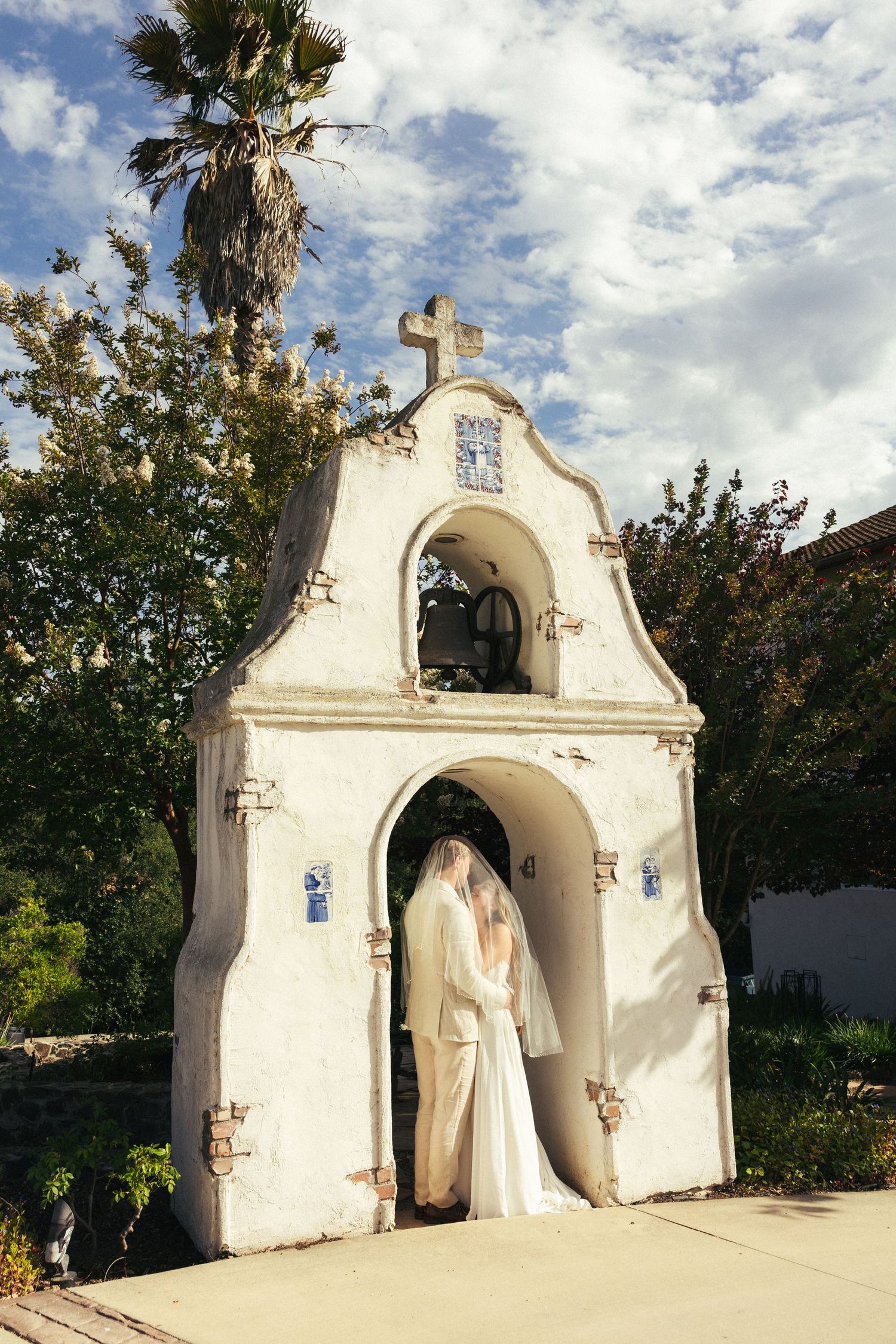 Bride in white gown and veil standing in arched doorway of Spanish mission chapel with cross and bell tower.