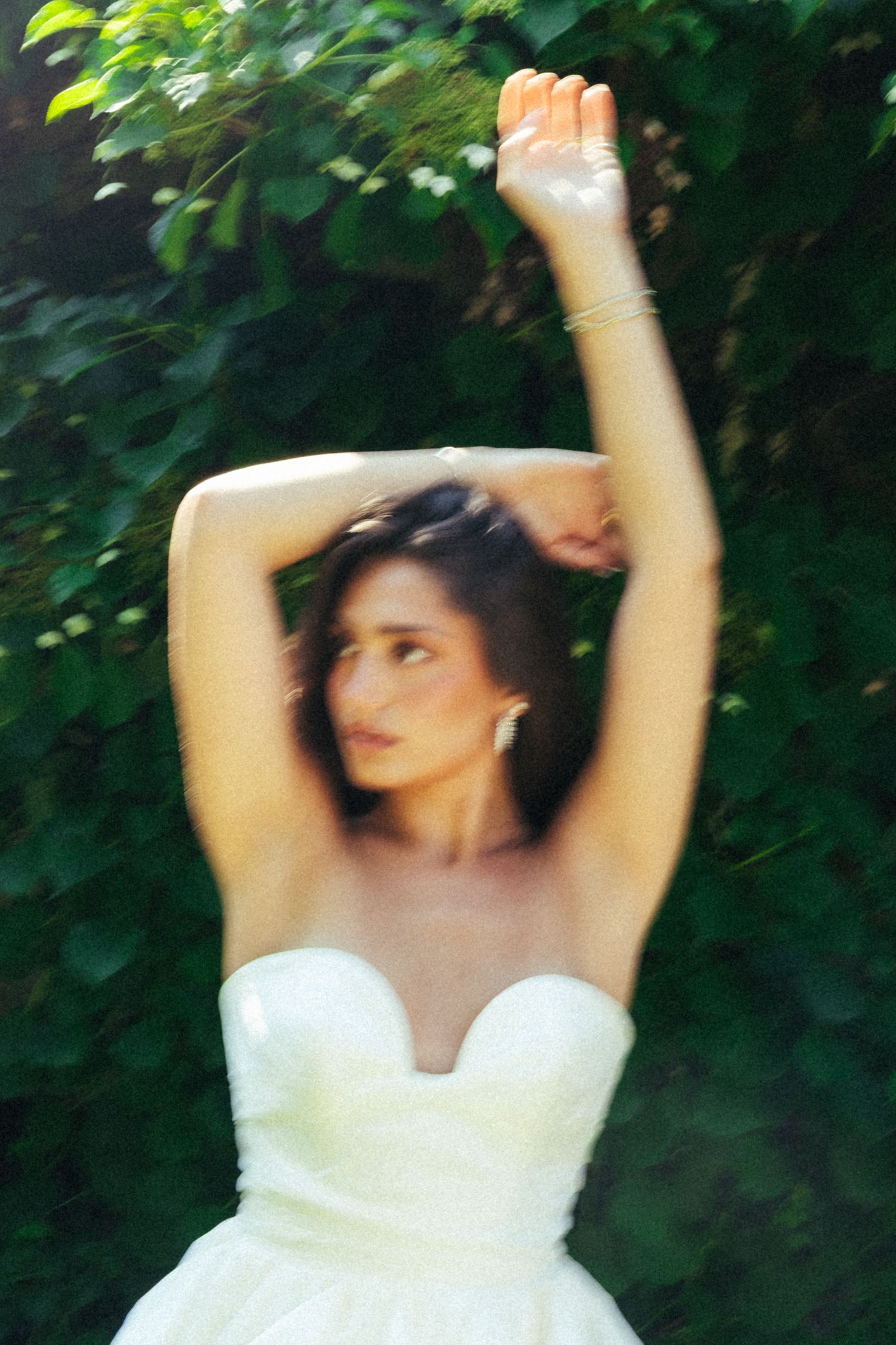 A woman in a white strapless gown raises her arms gracefully against a lush green leafy backdrop.