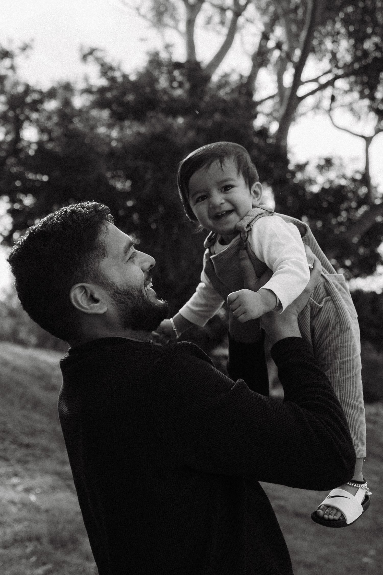 Black and white photo of a bearded father lifting his smiling baby boy outdoors near trees.