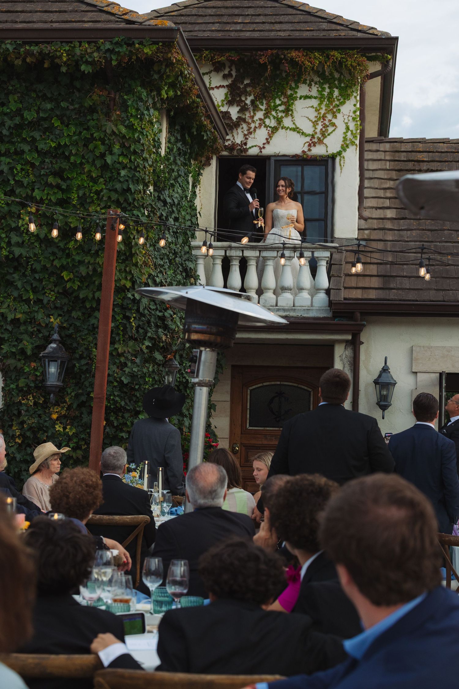 Bride and groom stand on ivy-covered venue balcony while wedding guests dine at outdoor reception tables below.