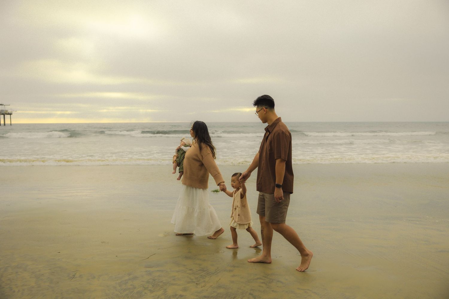 A couple walks along a misty beach shoreline at sunset while waves crash against the sand.
