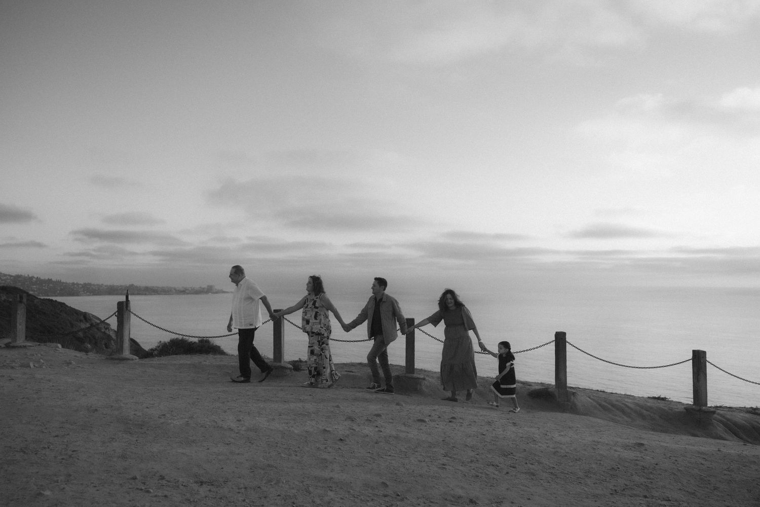 Silhouettes of people holding hands while walking along a coastal path at sunset with fence posts in view.