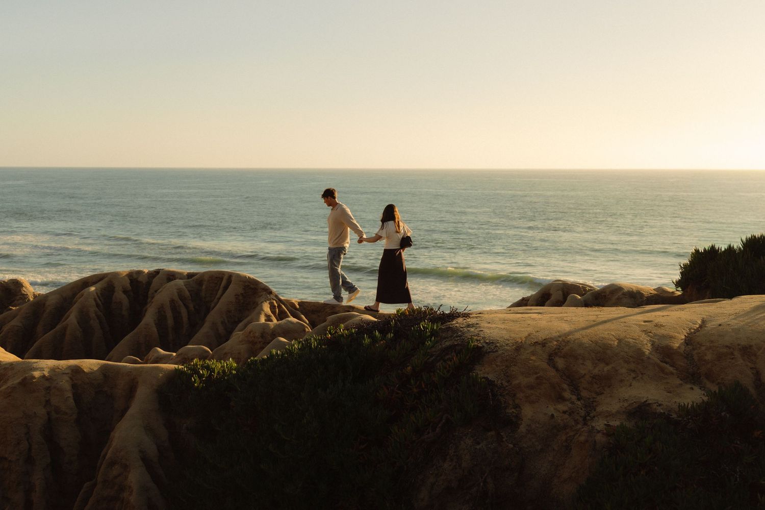 Silhouettes of people walking along a scenic coastal beach path at sunset with golden light reflecting on the ocean.