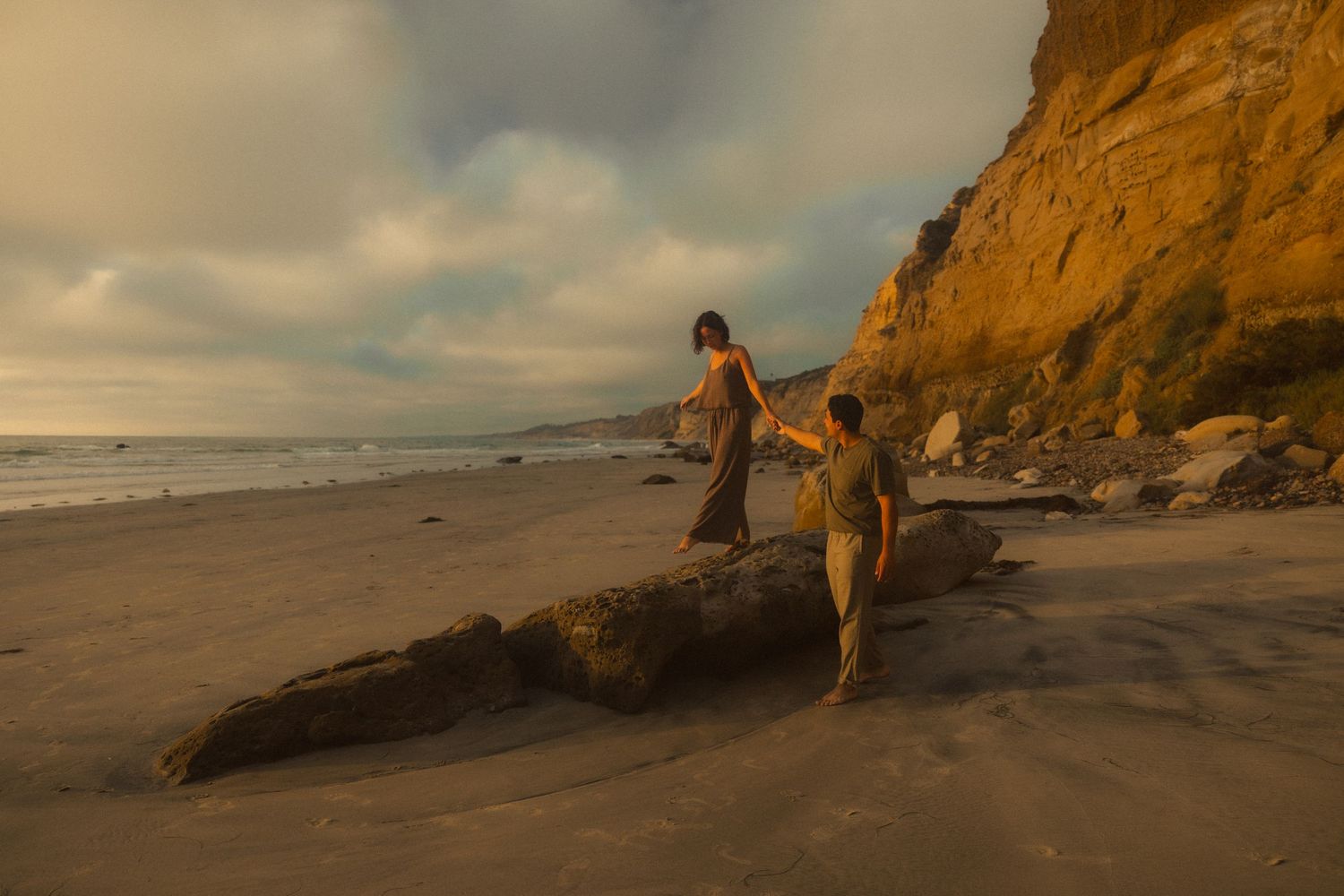 A person sits on coastal rocks at sunset while waves crash against the golden cliffs along a scenic beach.