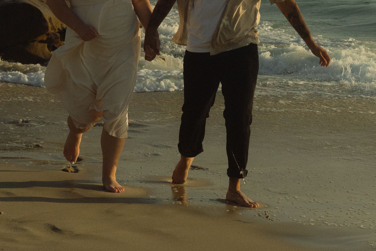 Legs of people walking barefoot along the shore with gentle ocean waves washing over the sandy beach.