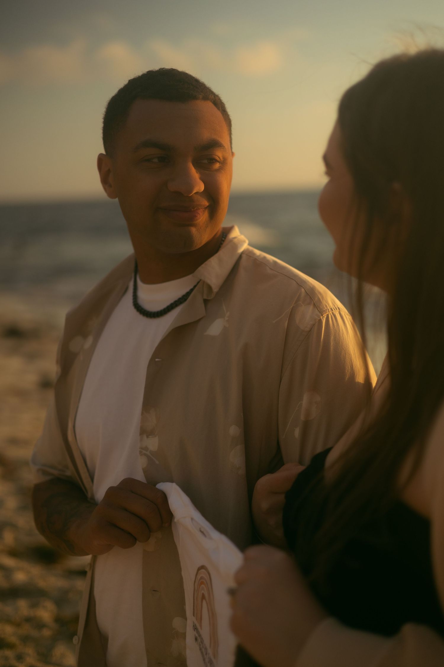 A couple shares an intimate moment at sunset along a beach shore in warm golden lighting.