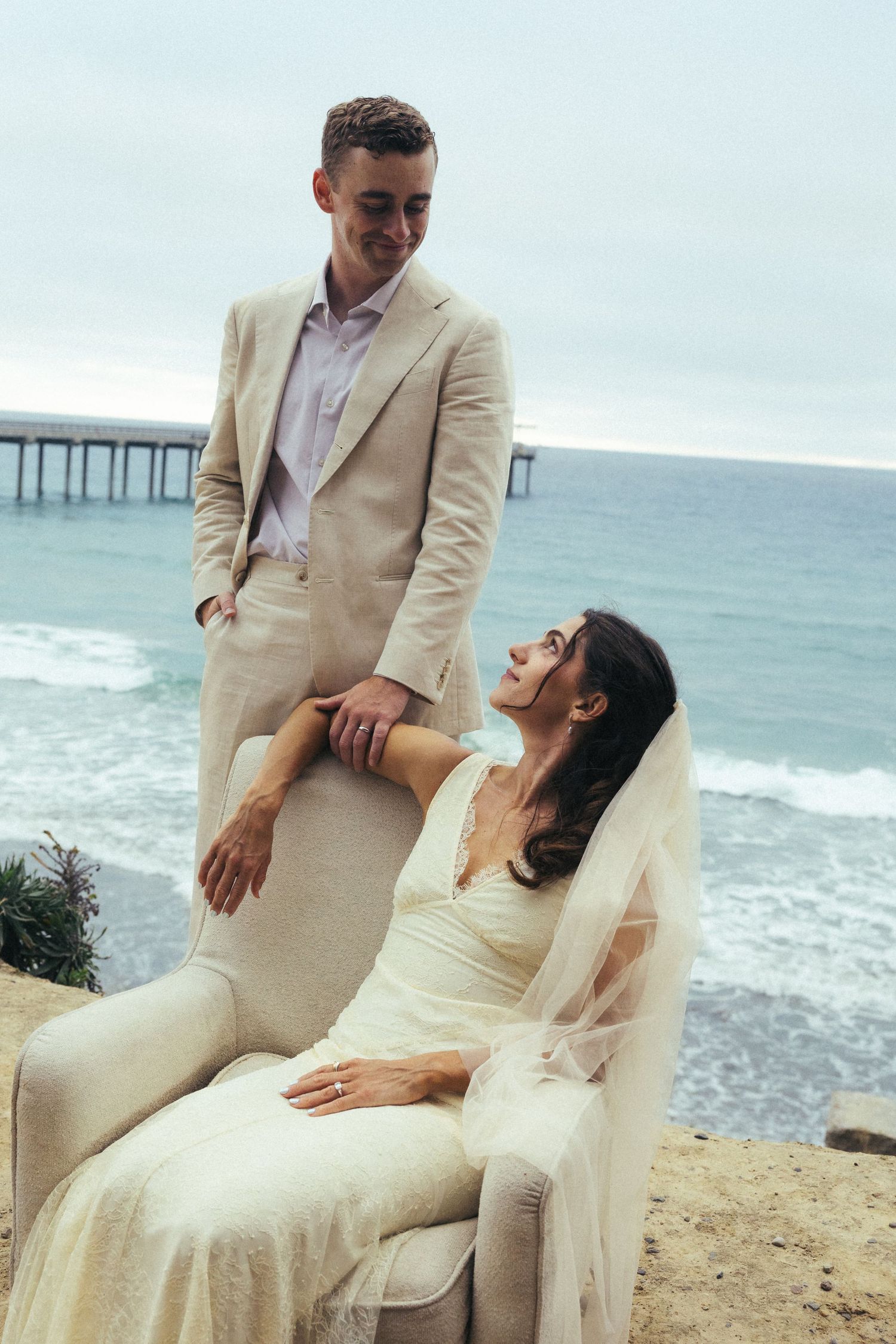 Couple in wedding attire posing by ocean pier, bride seated in chair smiling up at groom.