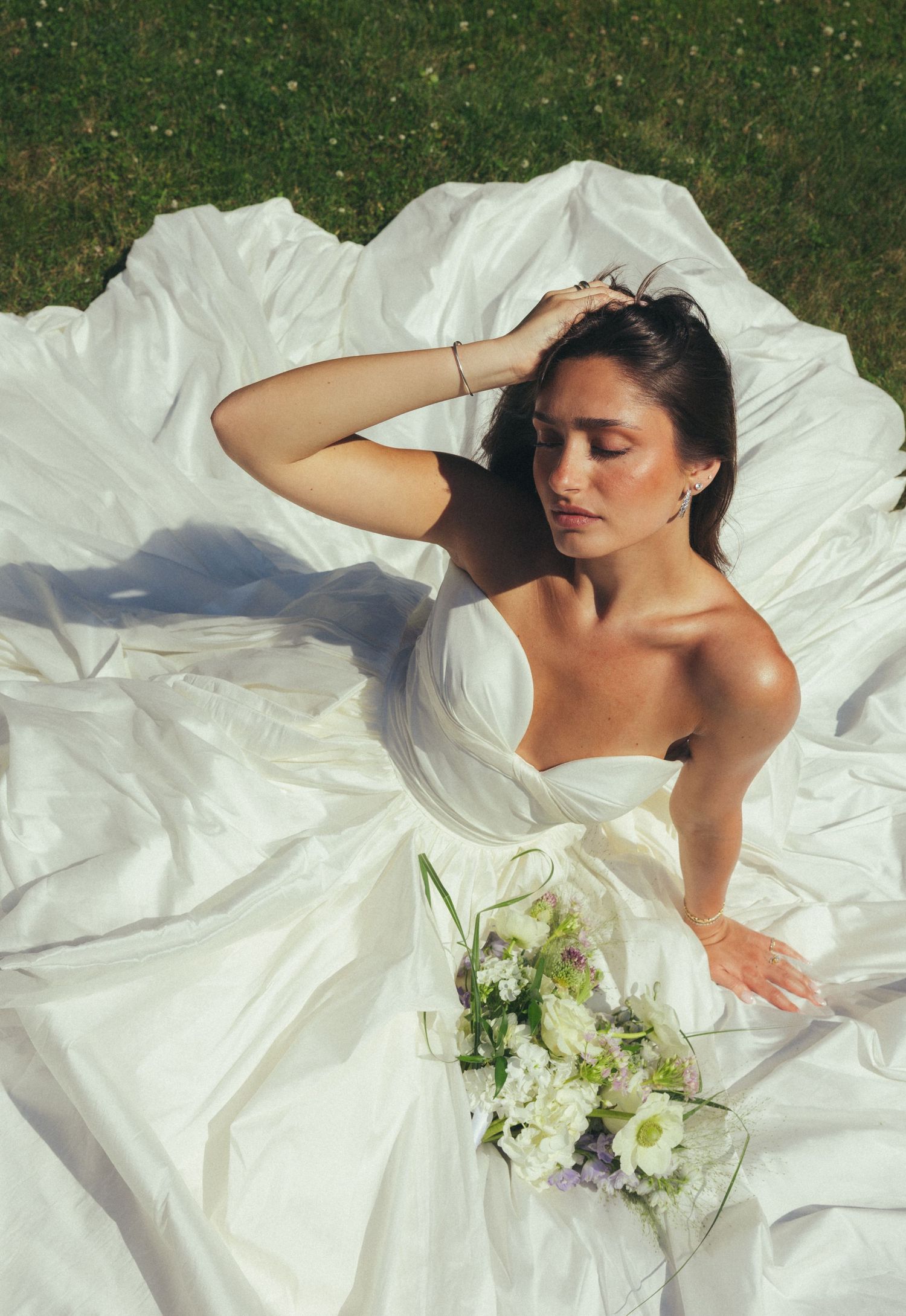 Bride in strapless white ball gown lying on grass holding bouquet, eyes closed in sunlight.