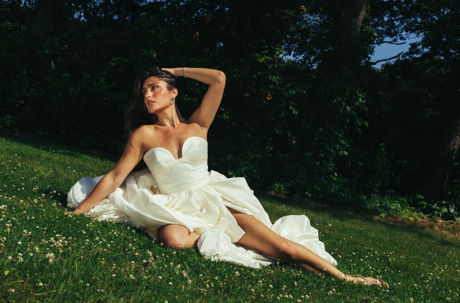 Woman in elegant white strapless gown posing barefoot on grass in golden sunlight.