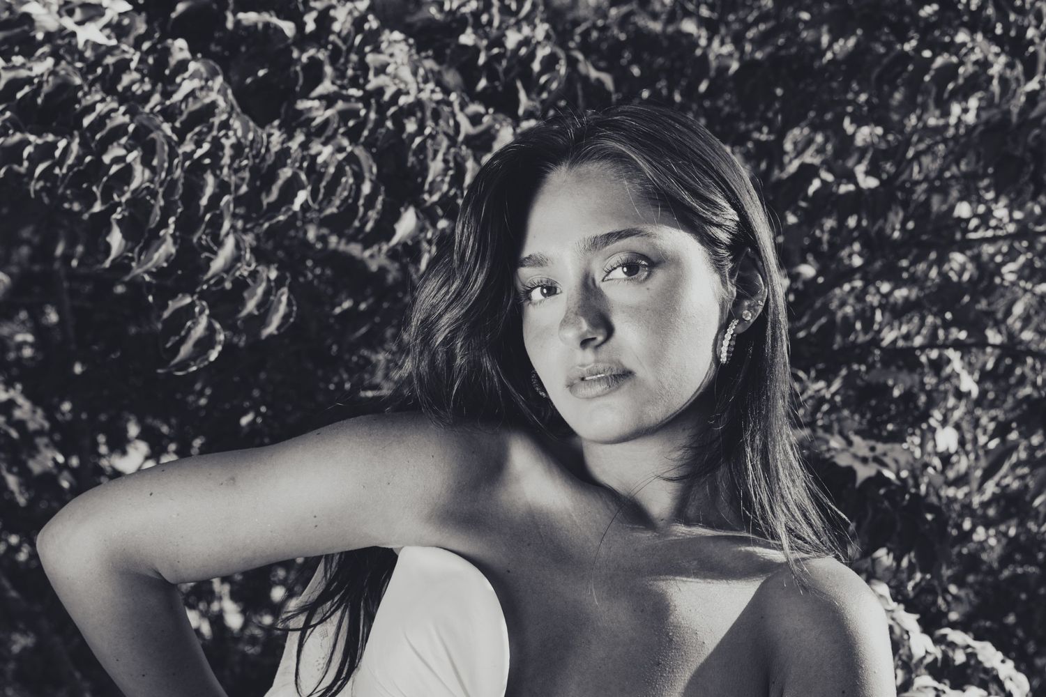 Black and white portrait of a young woman with long hair posing outdoors against lush foliage backdrop.