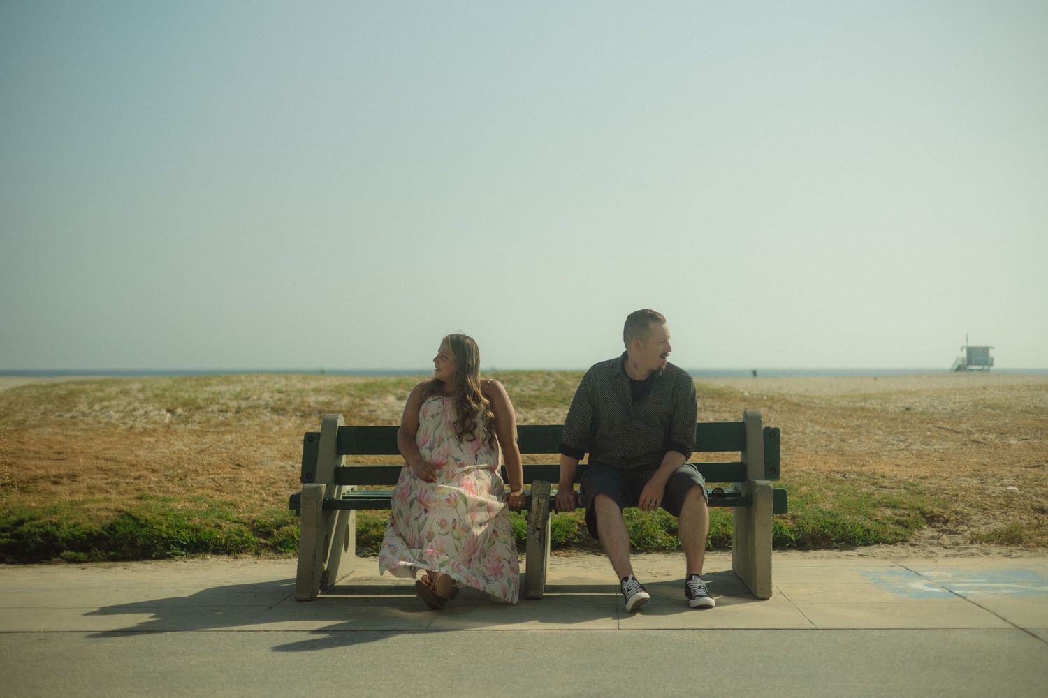 Two people sit on a wooden bench overlooking a barren coastal landscape on a hazy day.