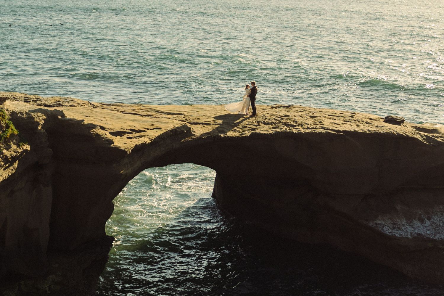 A silhouetted figure stands on a dramatic natural stone arch over the ocean during golden hour.
