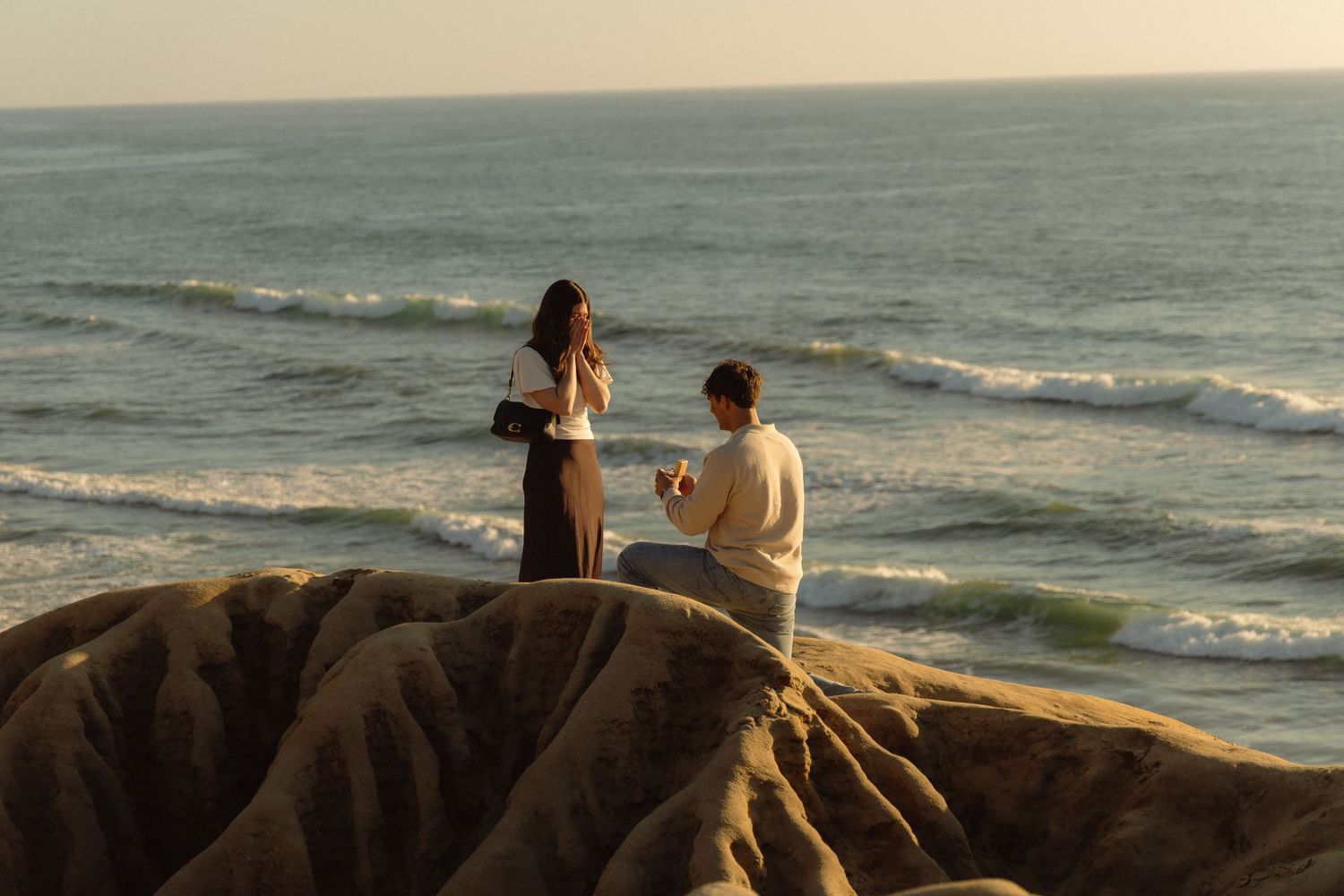 Couple sits together on rocky coastline watching waves crash against shore at sunset.