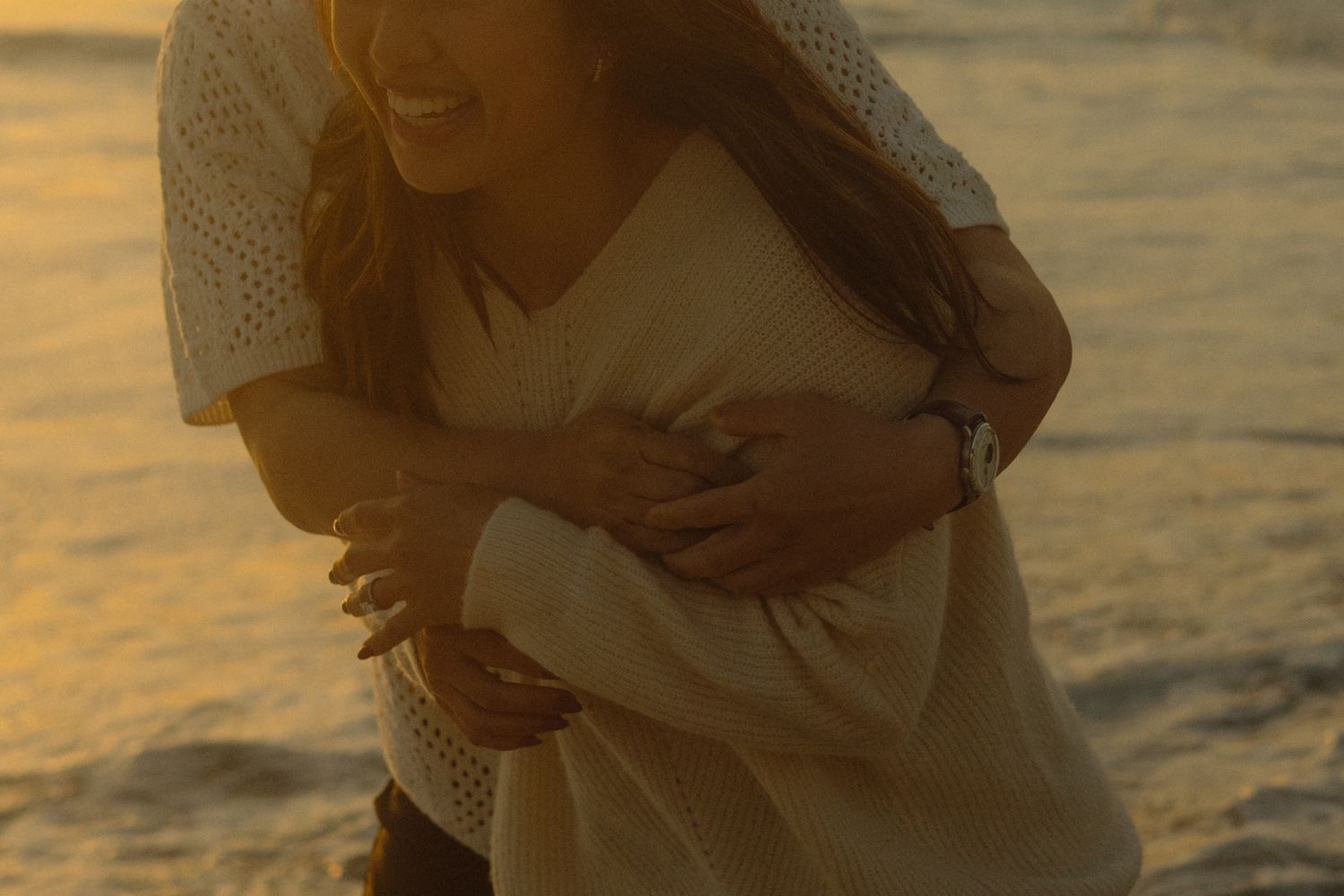 A couple embraces on the beach at sunset with golden light reflecting off the water in the background.