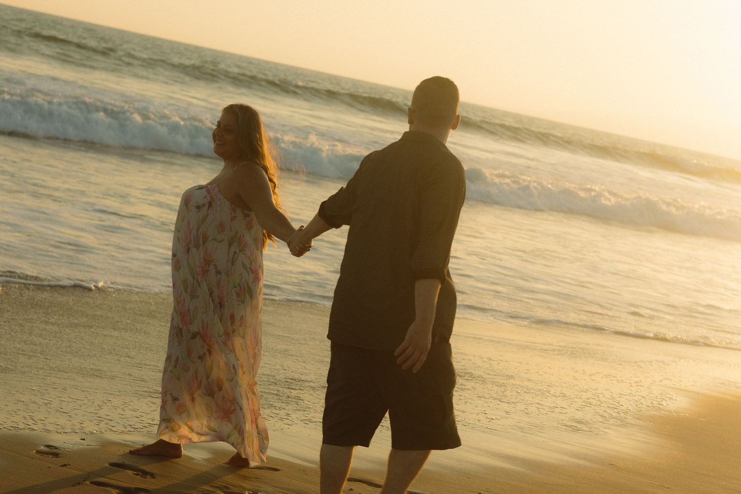 Two silhouettes hold hands while walking on a beach at sunset with ocean waves in the background.