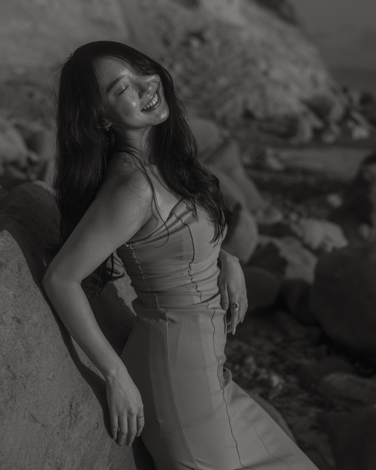 Black and white portrait of a person in elegant dress posing against rocky backdrop.