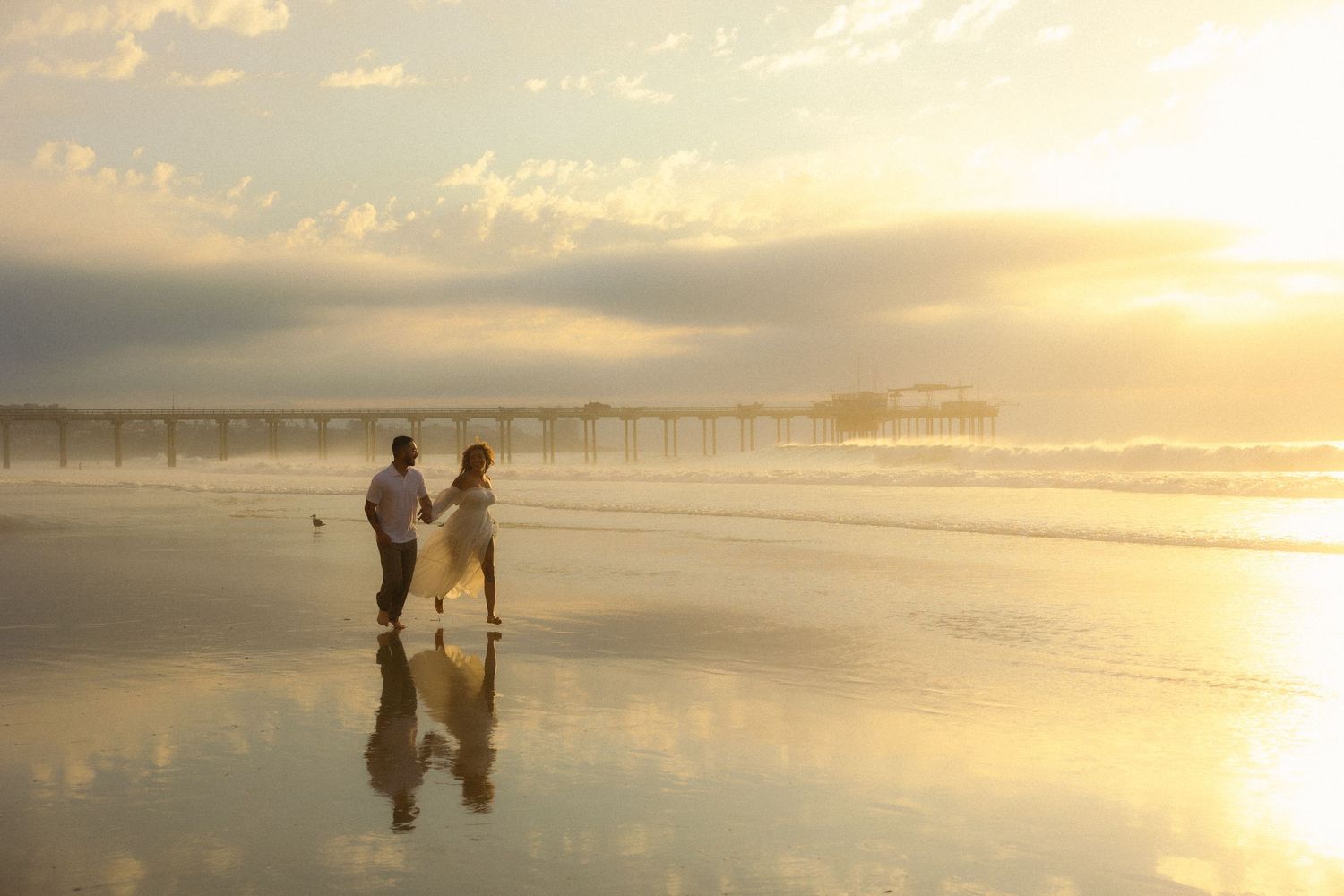 Silhouettes of people walking along a misty beach at sunset with a pier in the background.