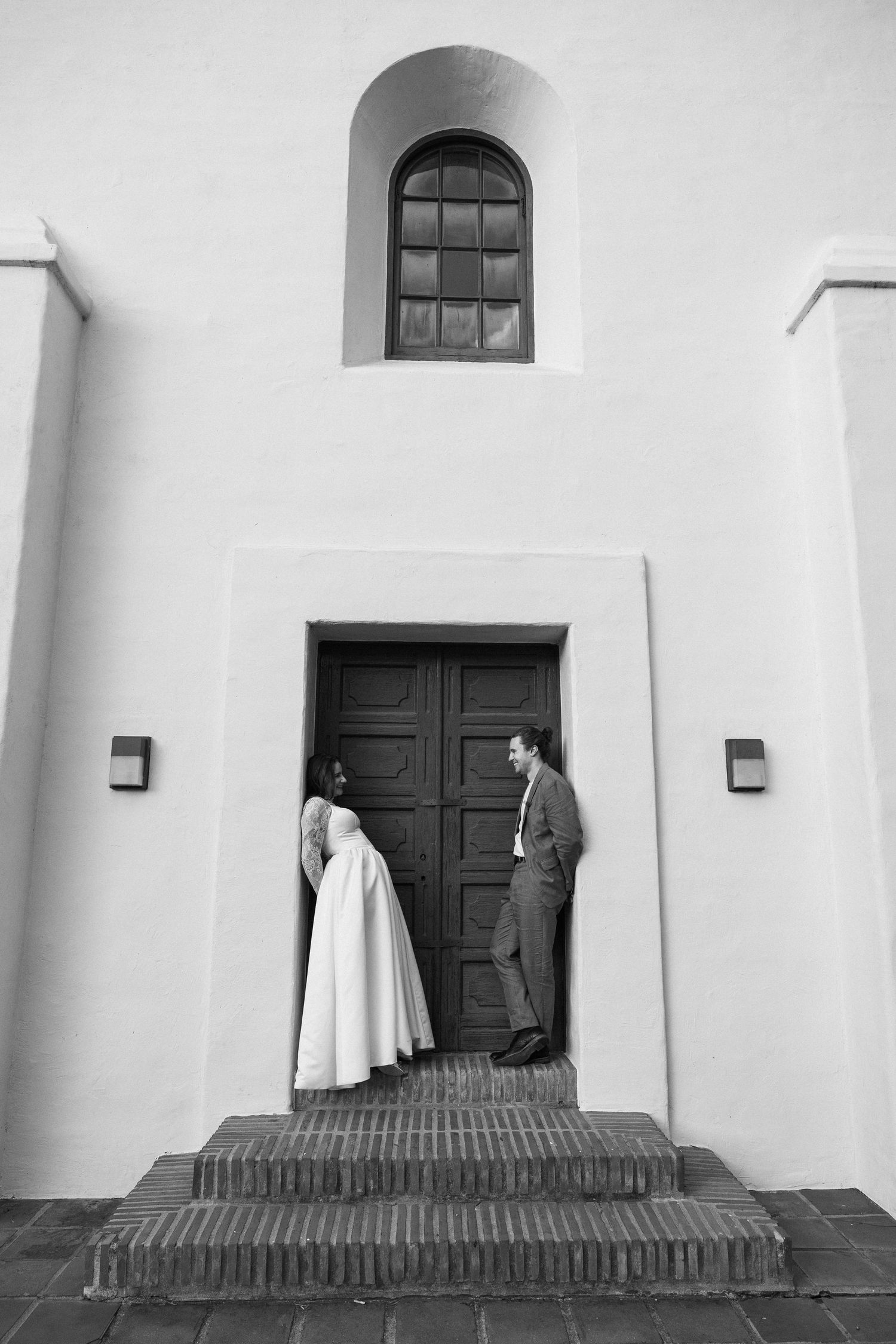 Black and white photo of bride and groom standing at church entrance doors on brick steps.