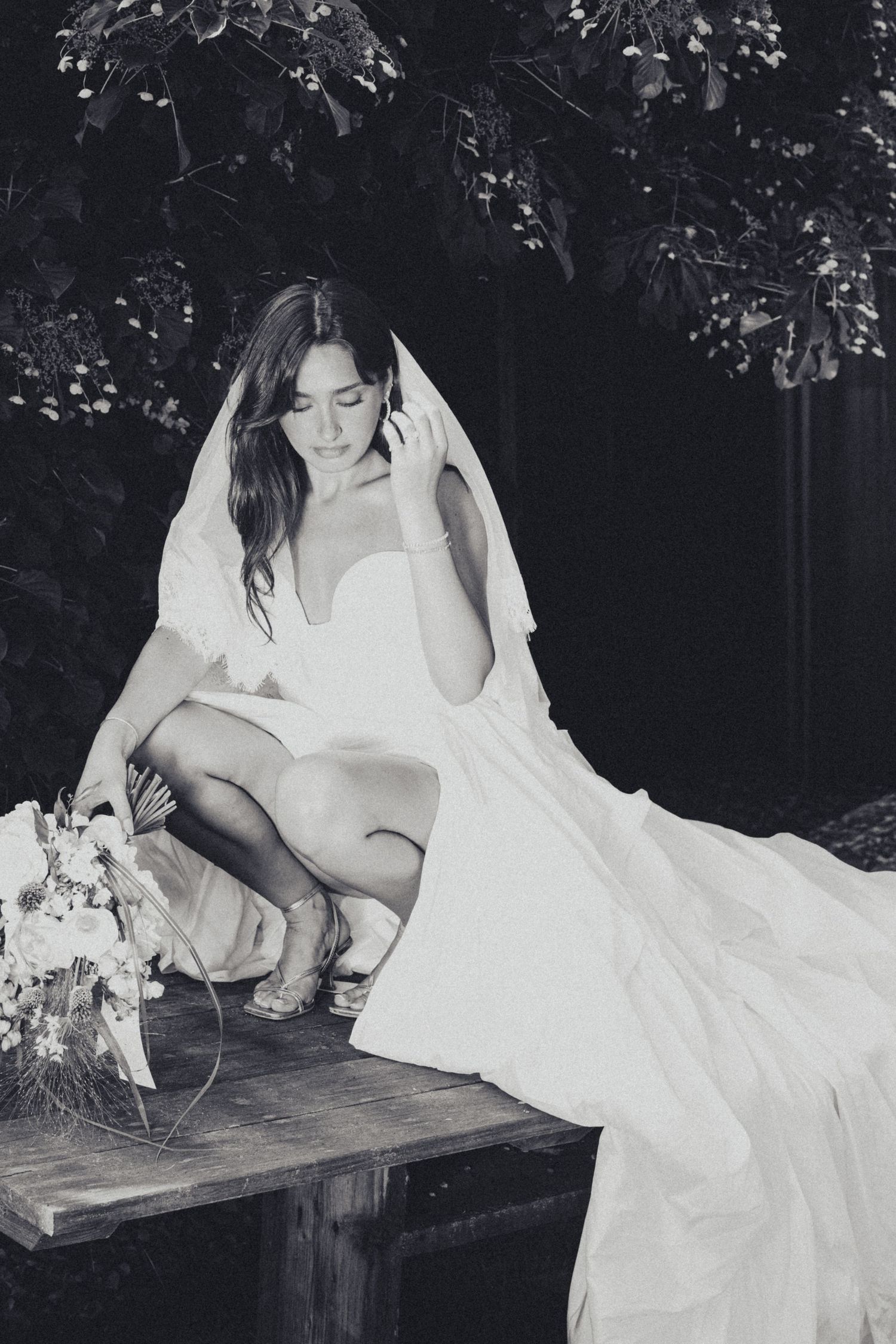 Black and white photo of bride in strapless gown crouching on bench with bouquet and veil.